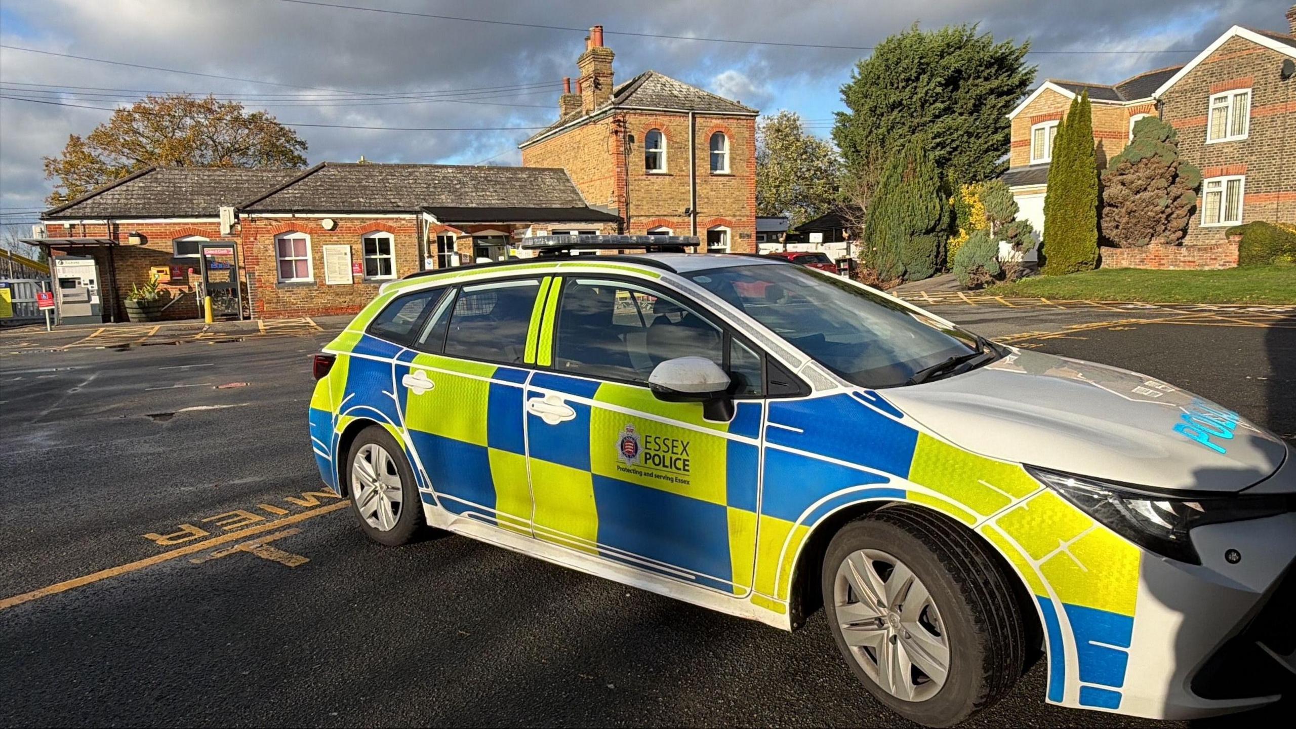 An Essex marked police car is parked in a car park with a redbrick railway station in the background