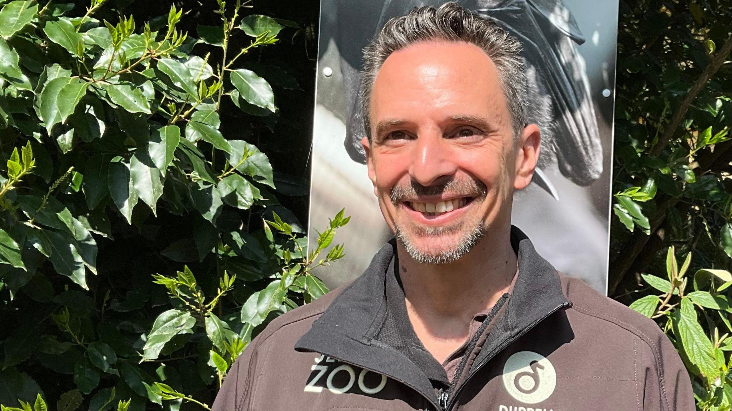 Matt Goetz is smiling at the camera. He is wearing a zipped sweatshirt with JERSY ZOO and DURRELL branding. He is standing outside and in front of a hedge and a sign which shows a photograph of a bat.
