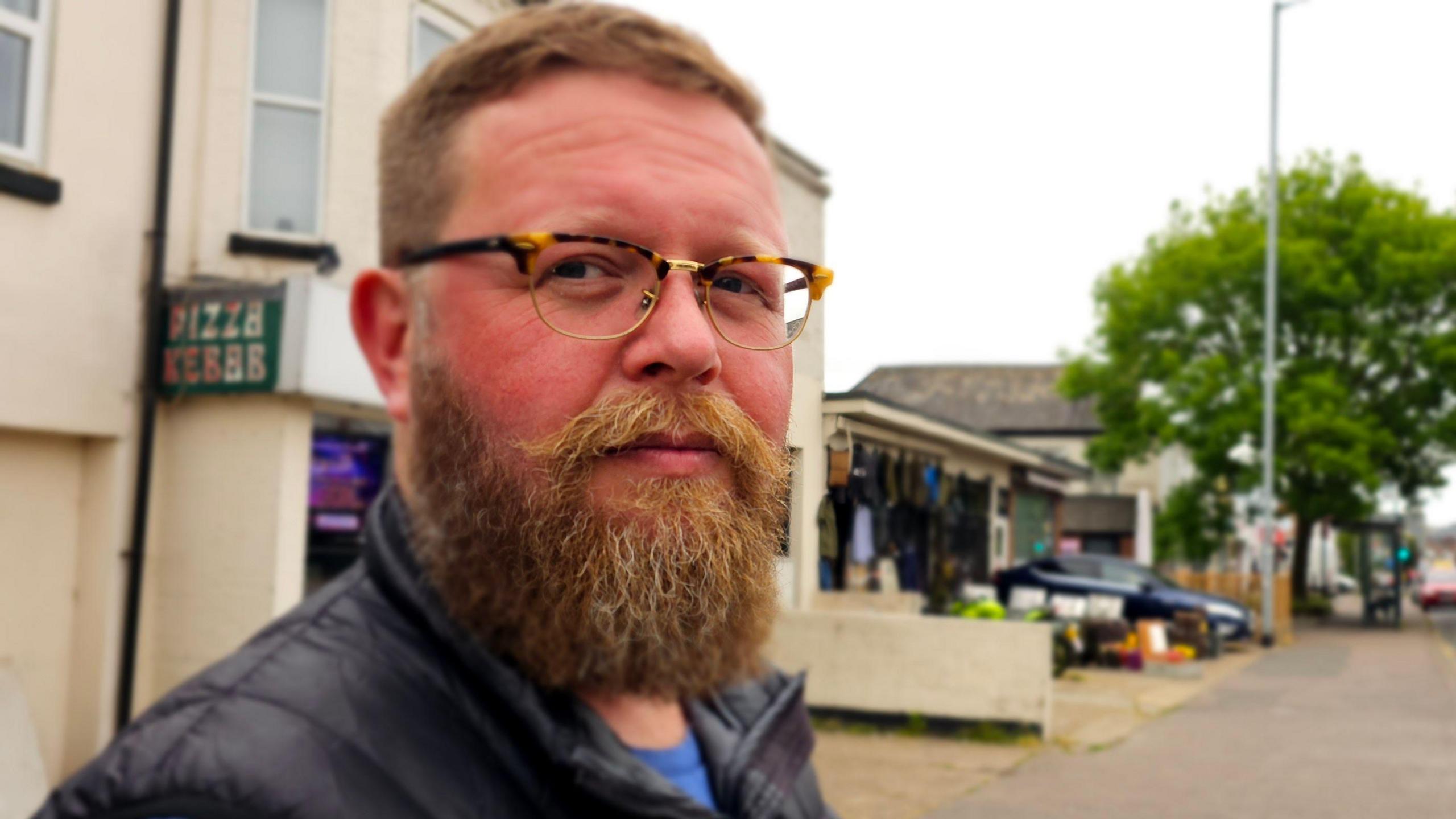 Liam Calvert is standing in a street in Norwich and looking at the camera. He is wearing glasses.