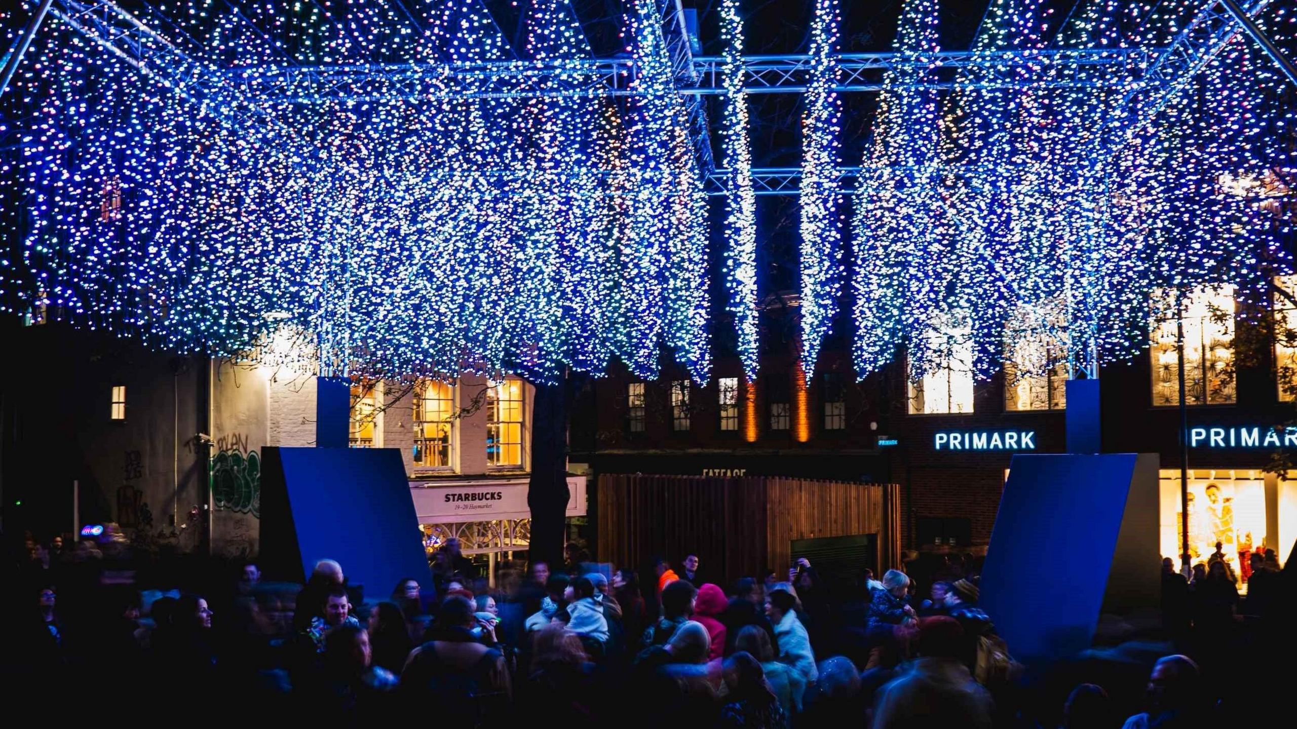 Crowds stand and look up to thousands of hanging Christmas lights, arranged in strips, which are blue, purple and green. The lights hang over an open street, next to a Primark store and near a Starbucks. 