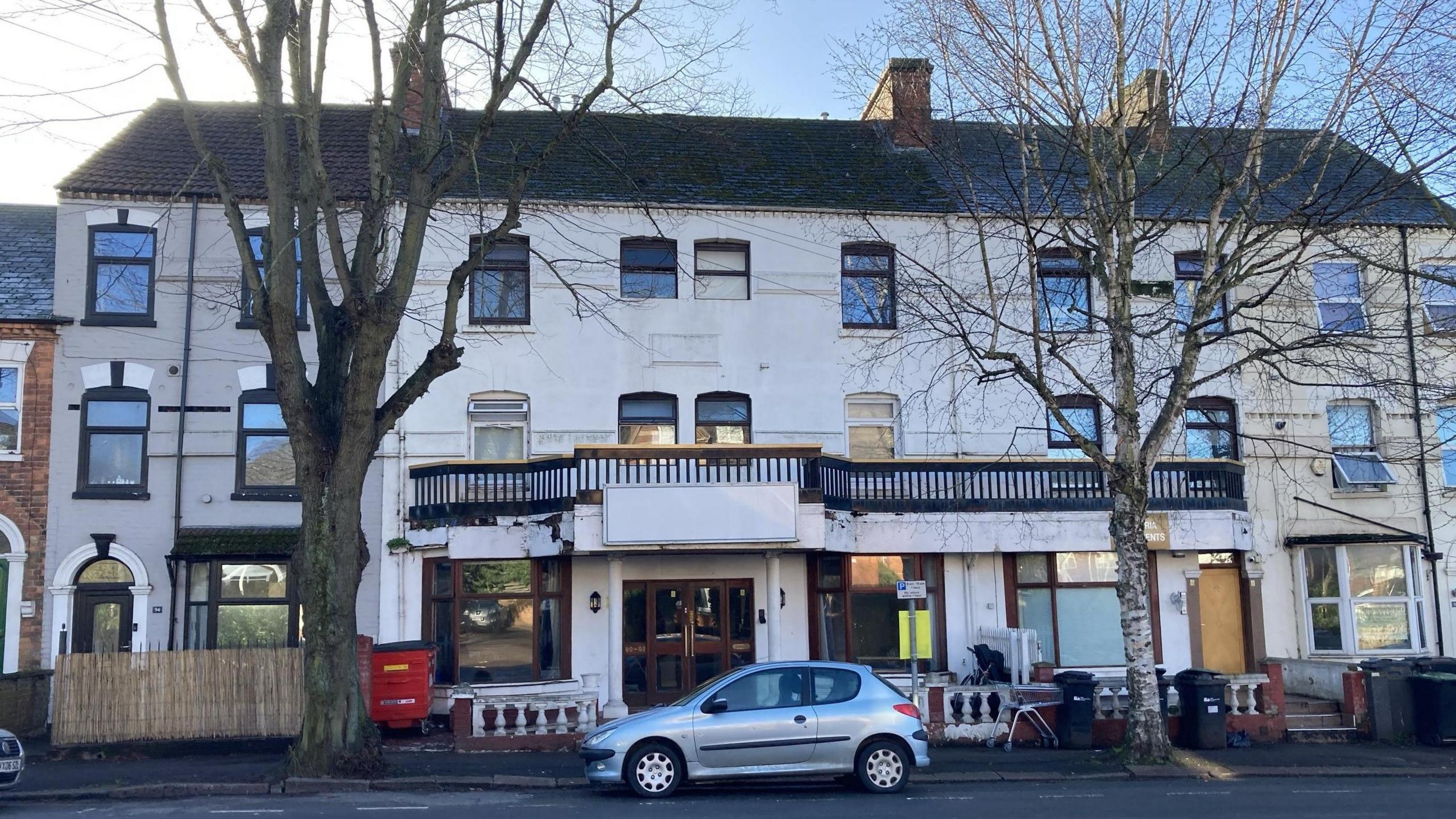 A white building with a long brown balcony on its second floor with many bay glass windows. A silver car is parked in front of the entrance. 