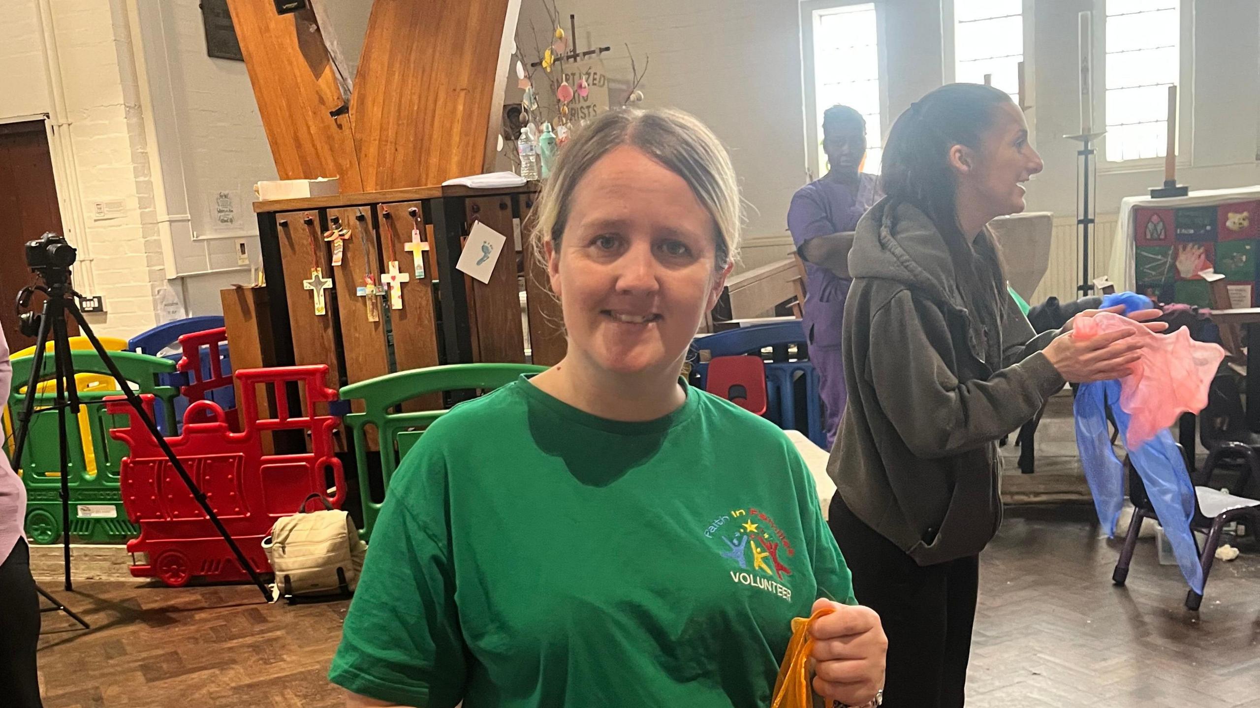 A blonde-haired woman in a green T-shirt stood in what looks like a church hall
