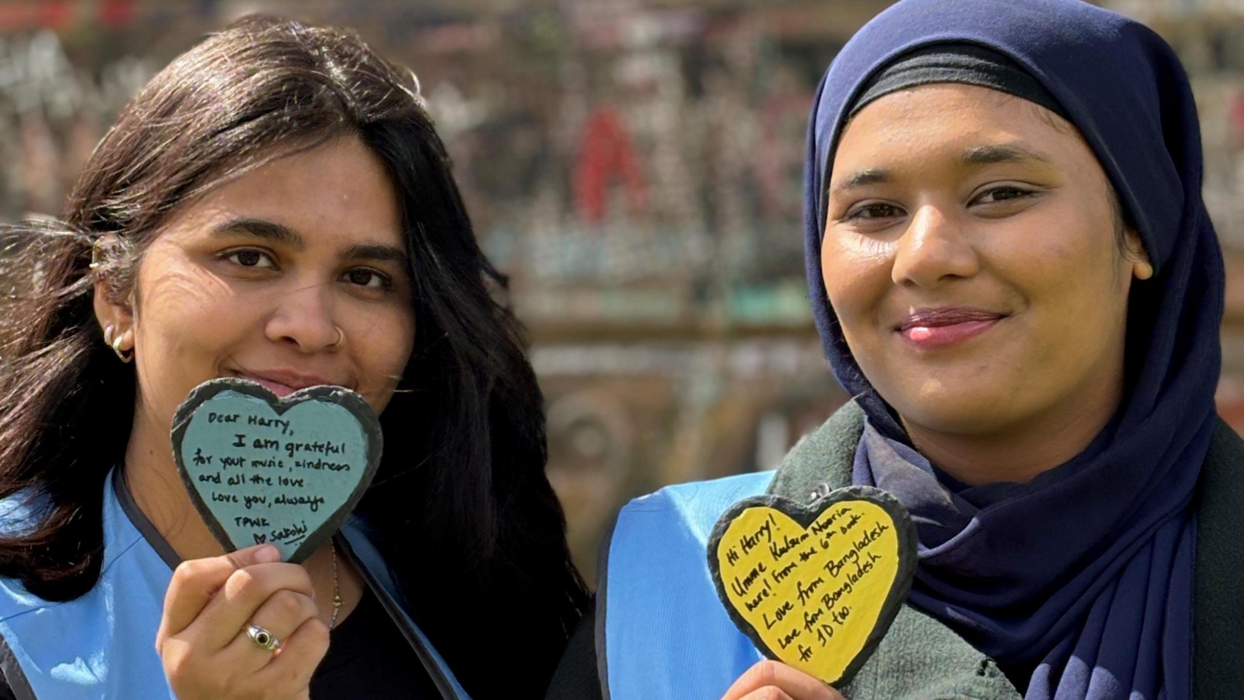 Two young women smile at the camera holding slate hearts with messages to Harry Styles