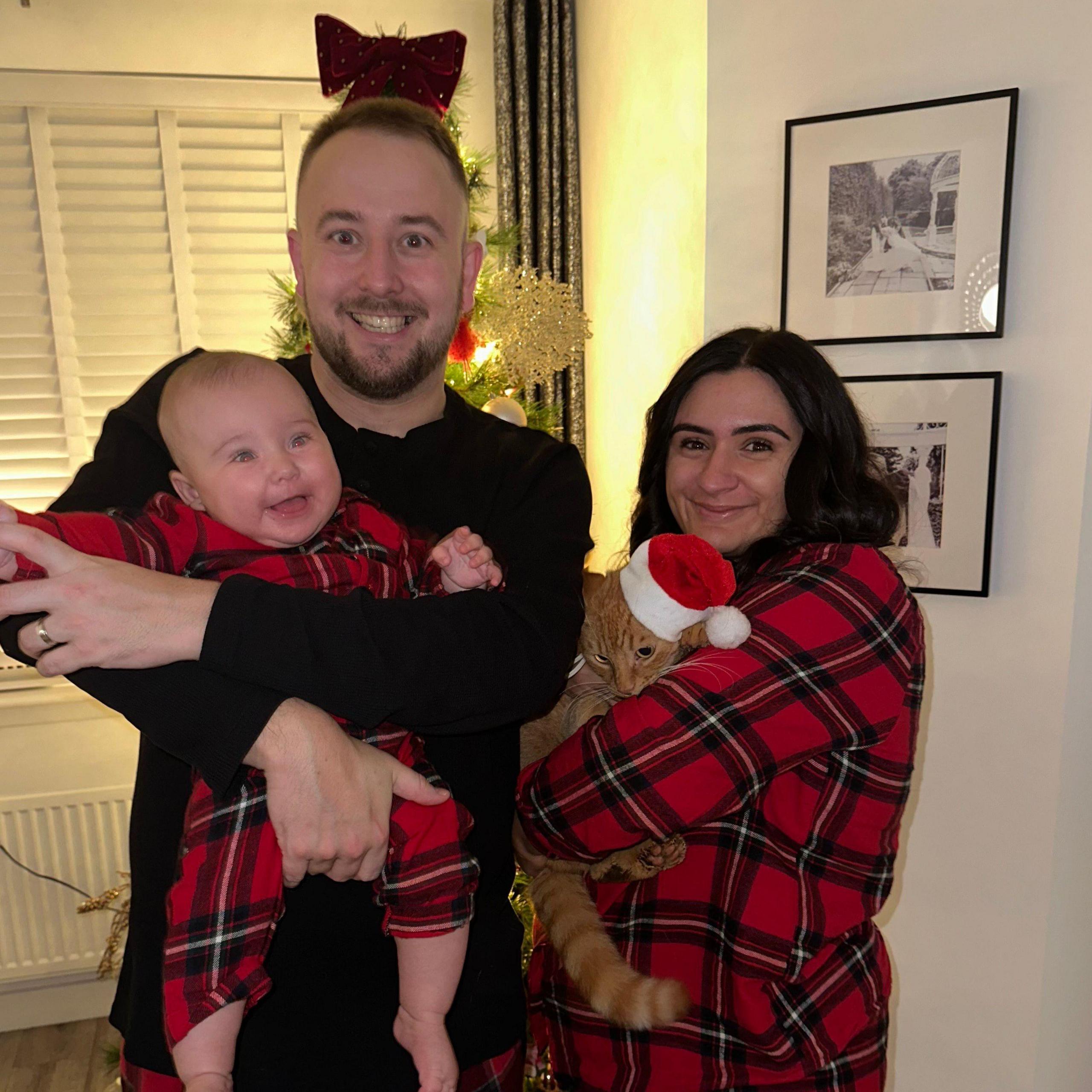 Aisha and David McCracken with baby Sophie at Christmas time. They are wearing matching red checked pyjamas and are standing in front of a Christmas tree. The baby is in her dad's arms and is a few months old. Aisha is holding a ginger cat that is wearing a Christmas hat.