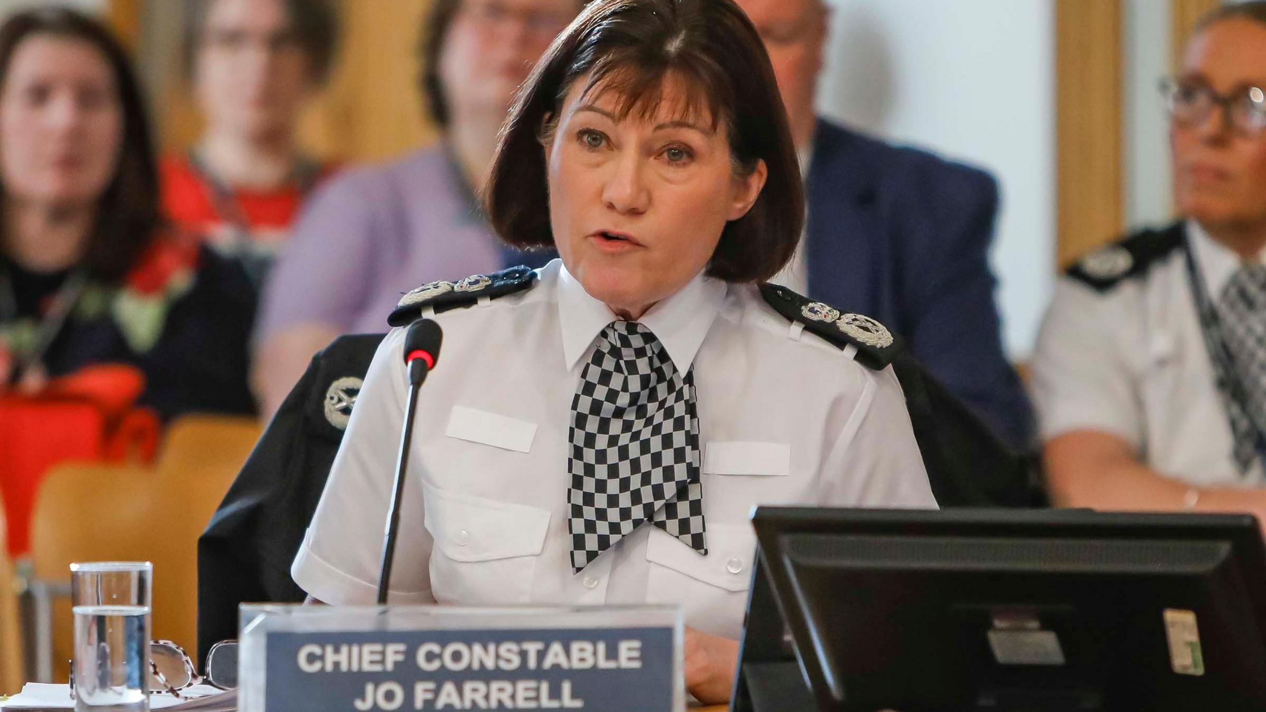 Jo Farrell, who has short brown hair, wearing a white police uniform with black badges sewn on the shoulders speaks into a microphone as people site behind her in the public gallery. There is a sign on the desk in front of her with her name and rank