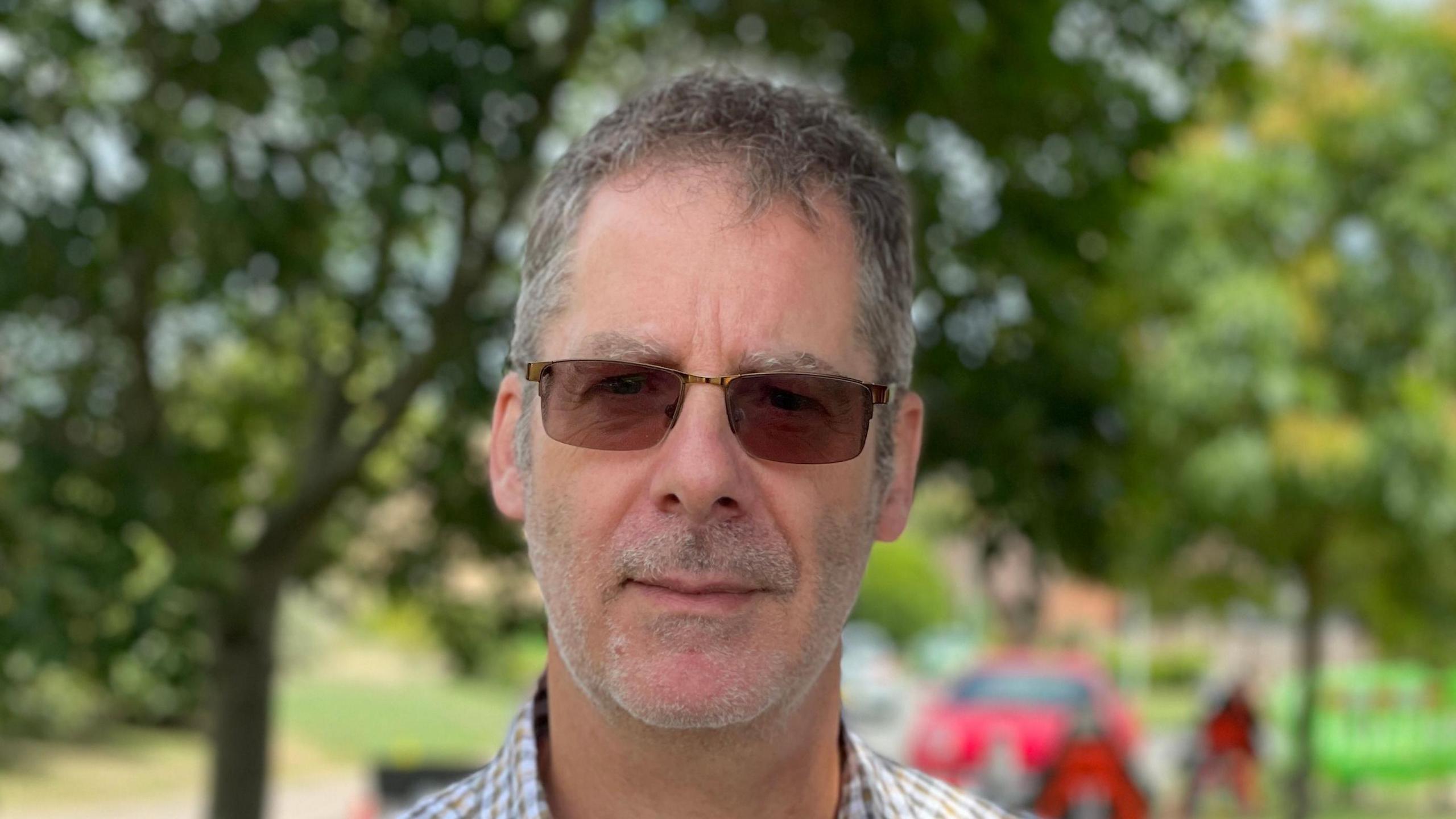 Mal Bridgeman smiles at the camera. He has short grey hair, wears sunglasses and a chequered shirt. 