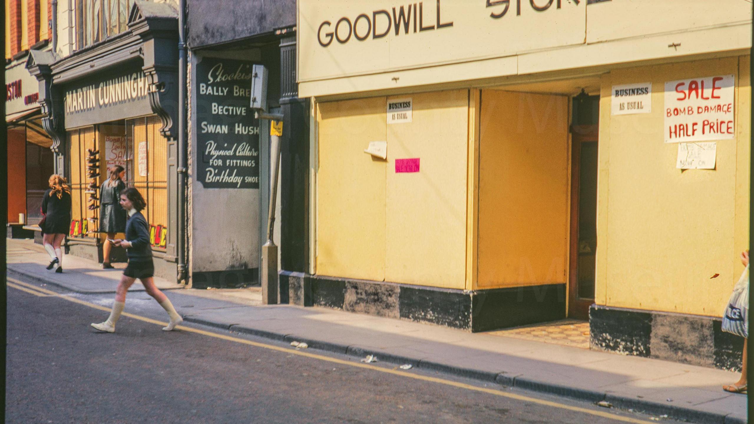 A digitalised photo from a film. It is of a yellow shop front on a street.  Shops visible are Emerson's grocery, Martin Cunningham and Goodwill store. A sign on the board reads, "BUSINESS AS USUAL SALE BOMB DAMAGE HALF PRICE"