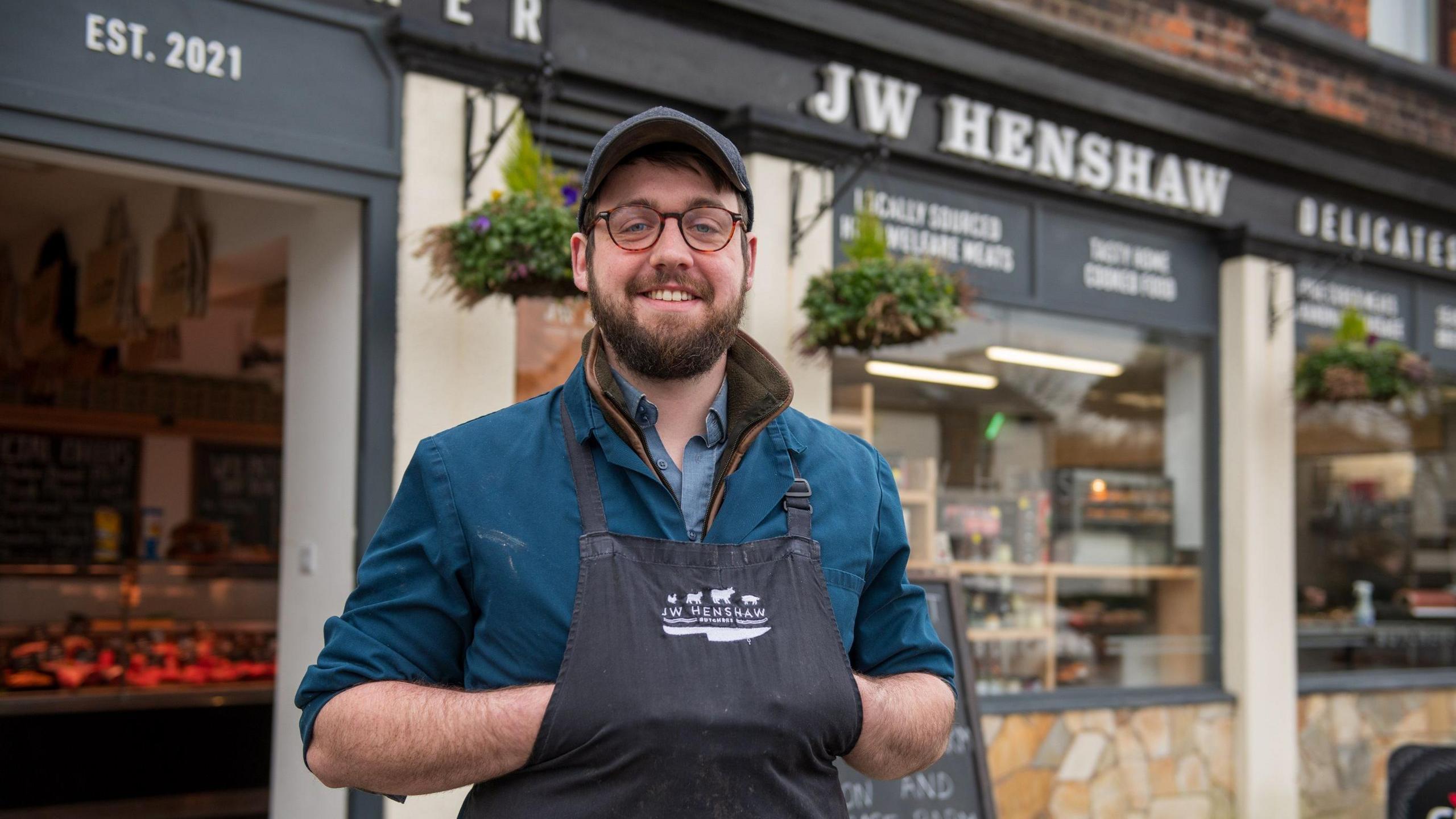 James Henshaw wearing a dark baseball cap and butcher's apron, smiling outside his business front