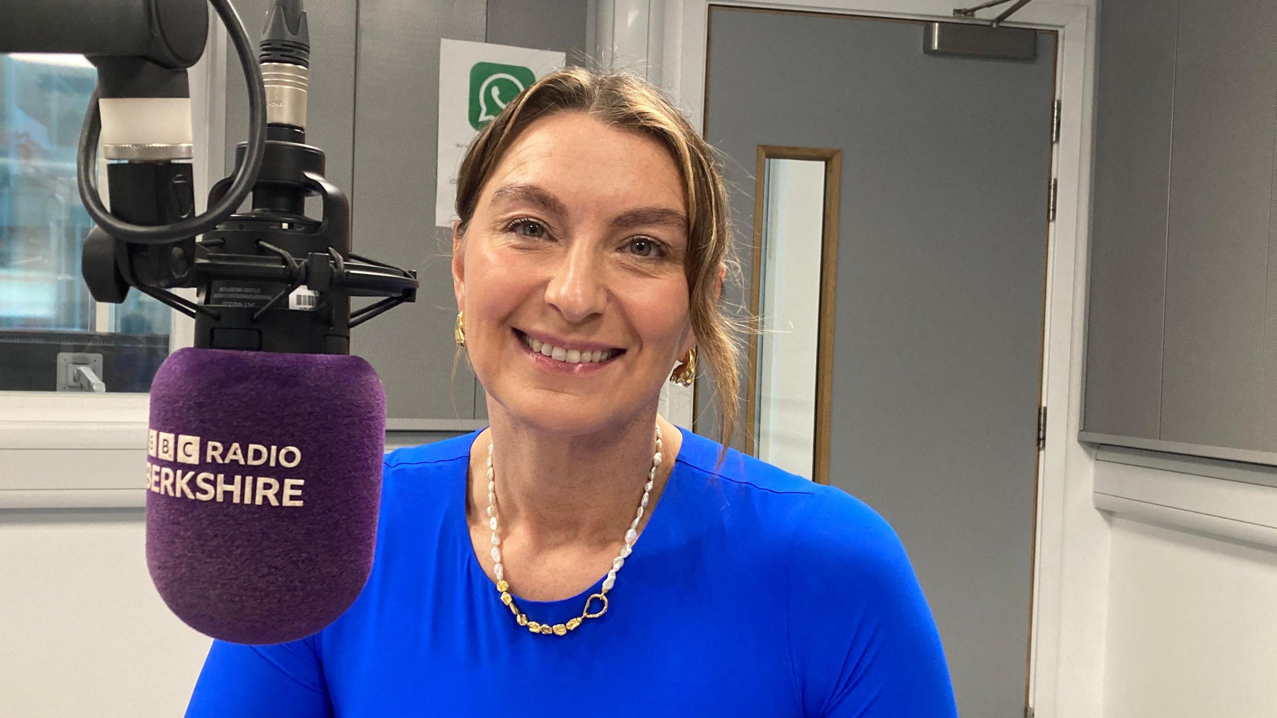 Rachel de Caux sits in front of a BBC Radio Berkshire microphone in a bright blue shirt and a golden and white necklace.