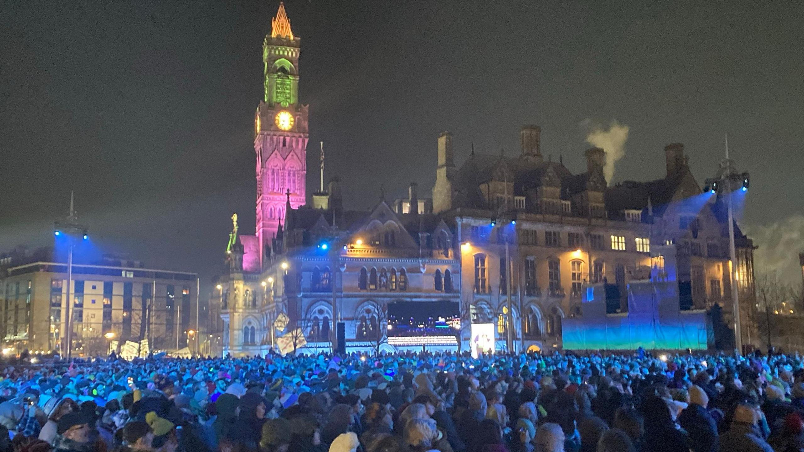 Building lit up with colours as thousands of people stand bathed in light watching a show below