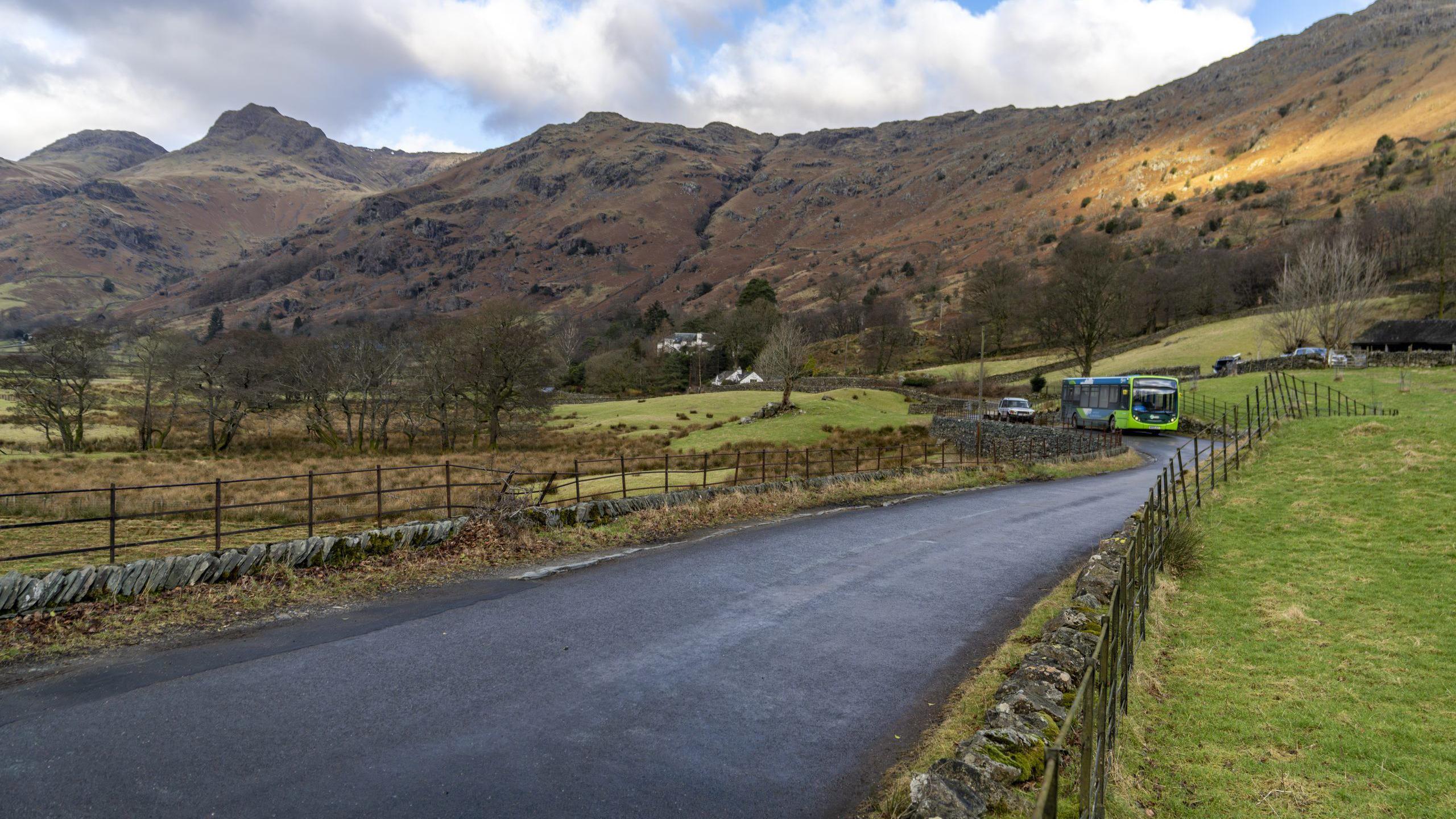 A bus pictured in the Lake District. The bus is coming round a corner and the fells are visible in the background.