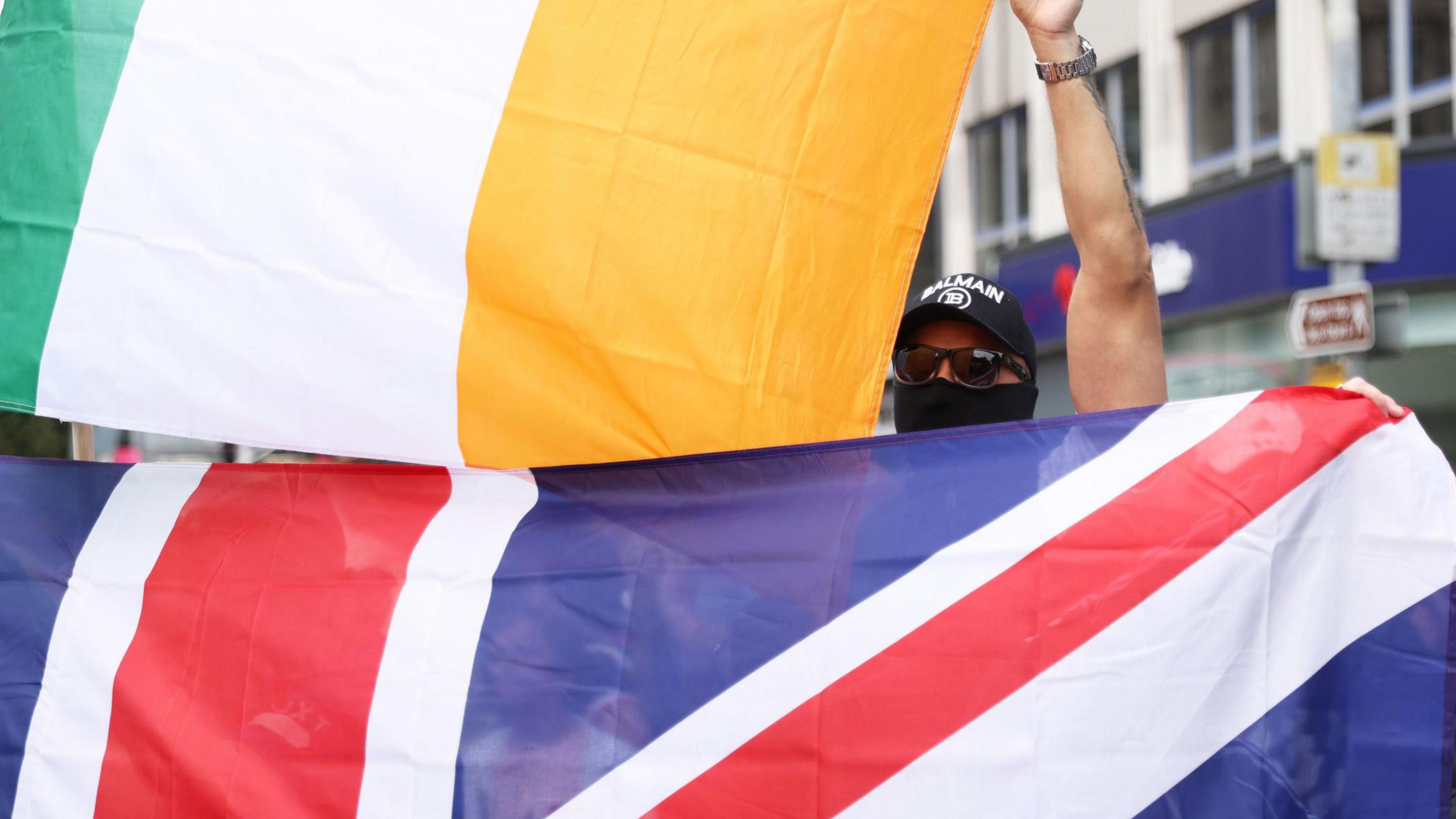 A Union flag and Irish tricolour held side by side. A man wearing a hat, face covering and sunglasses holds the tricolour.