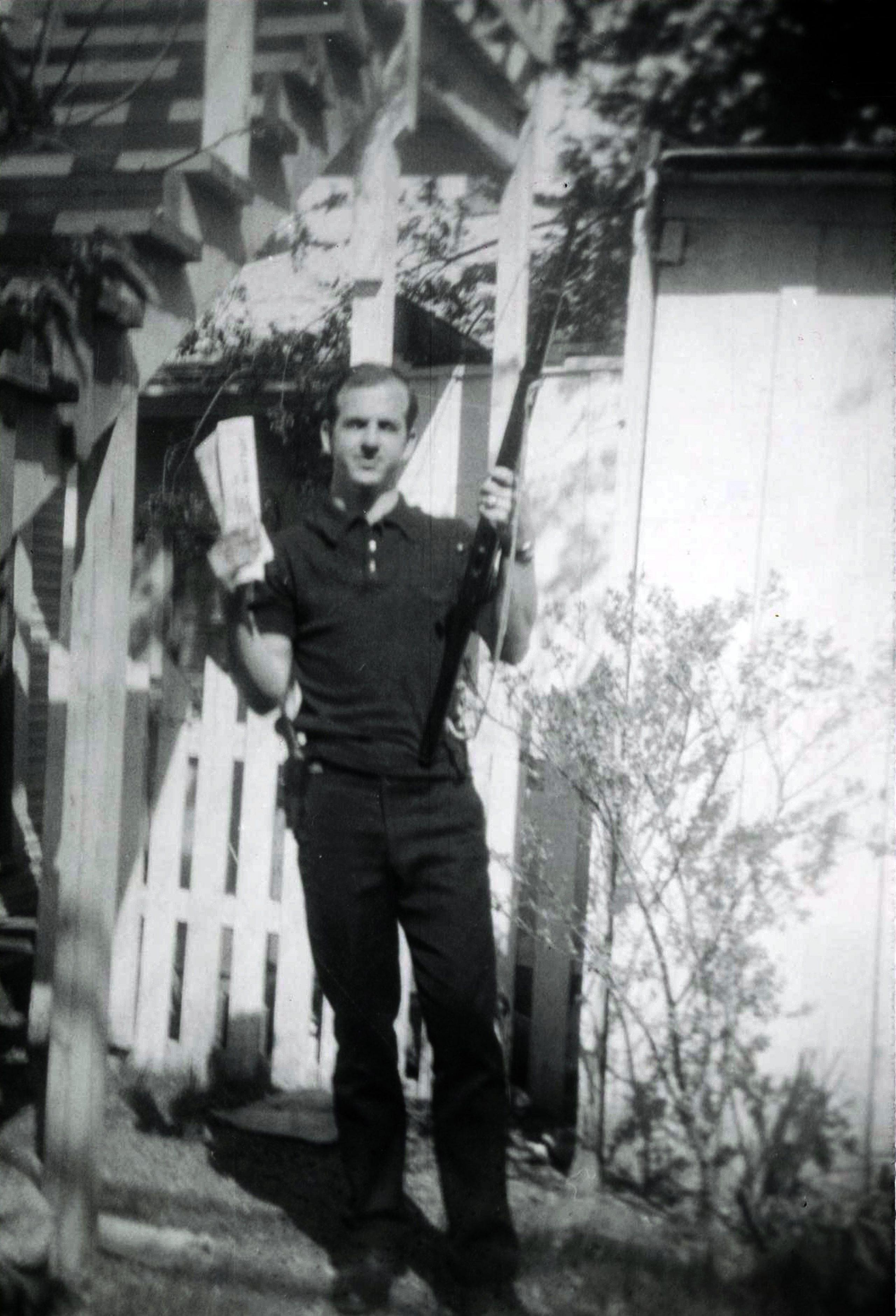 A black and white and slightly blurry image of Oswald holding a gun and papers, taken from a distance, with a fence and stairs in the background