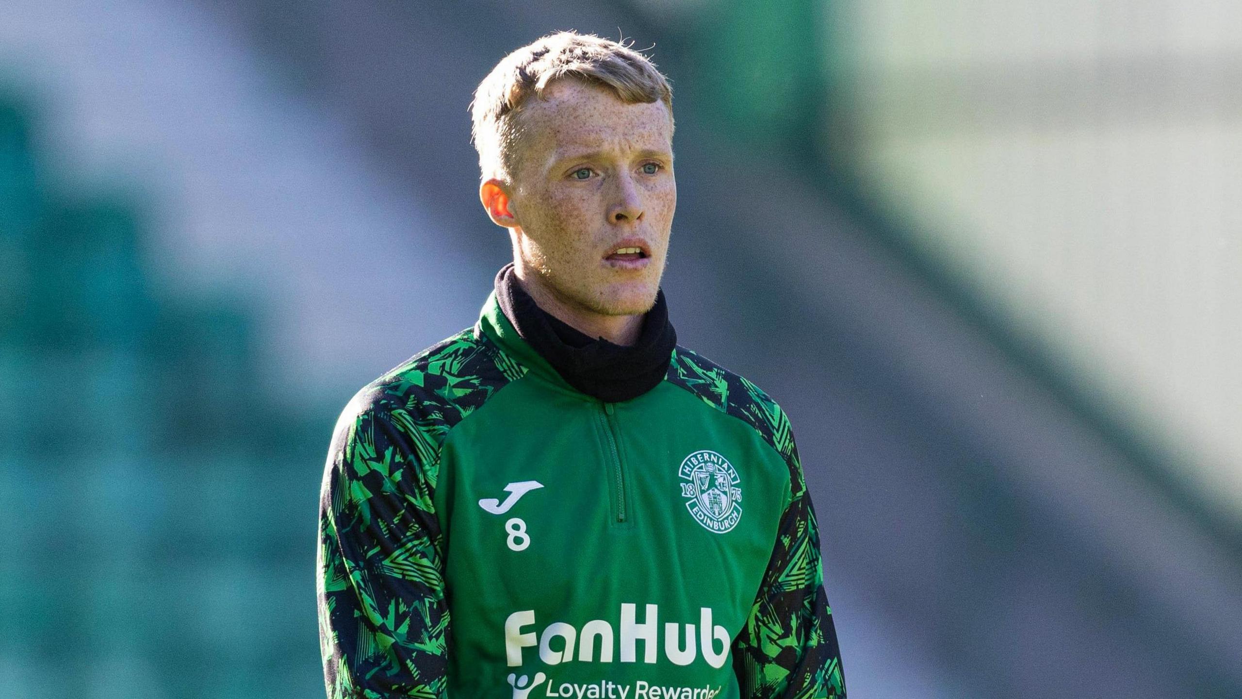Jake Doyle-Hayes wearing Hibs training gear. He is wearing a green top with the Hibs badge in the top right corner. He is wearing long green and black sleeves and black gloves. He has fair hair and is looking slightly off camera.