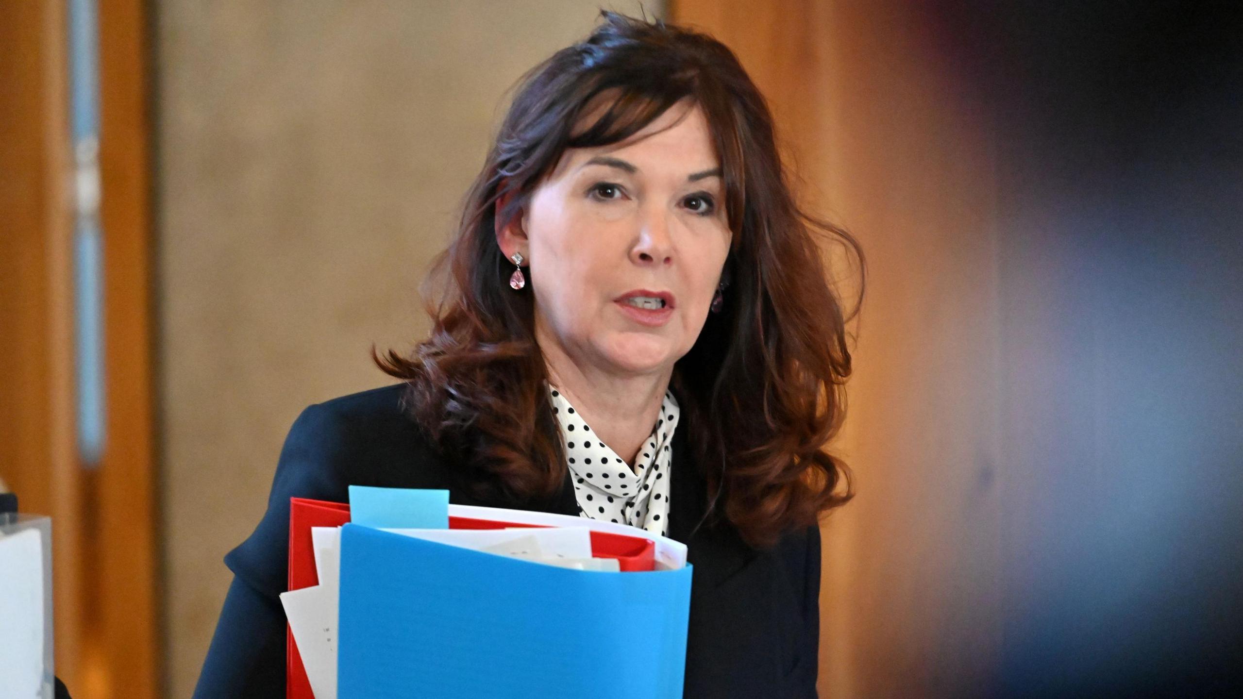 Dorothy Bain has long brown hair. She is wearing a suit jacket over a white blouse with black dots on it. She is carrying a folder with papers in it.