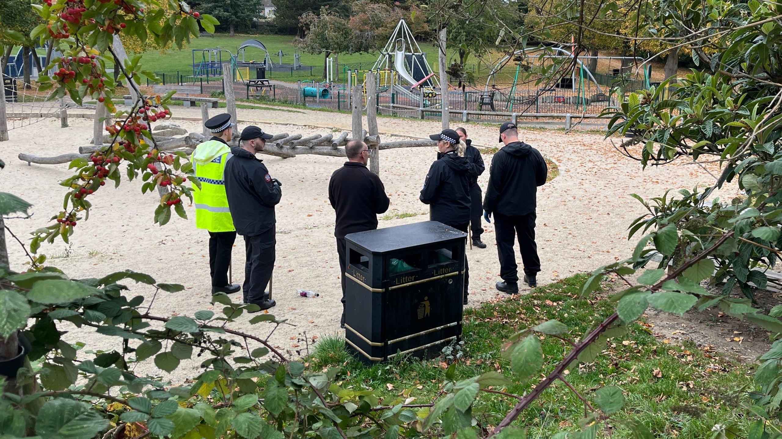 Five uniformed police officers stand in a children's outdoor playground. A black litter bin is in the near foreground. The floor of the park is made of sand. The officer on the left of the group wears a police hat and a fluorescent yellow jacket. There are some green leafy trees in the park. 