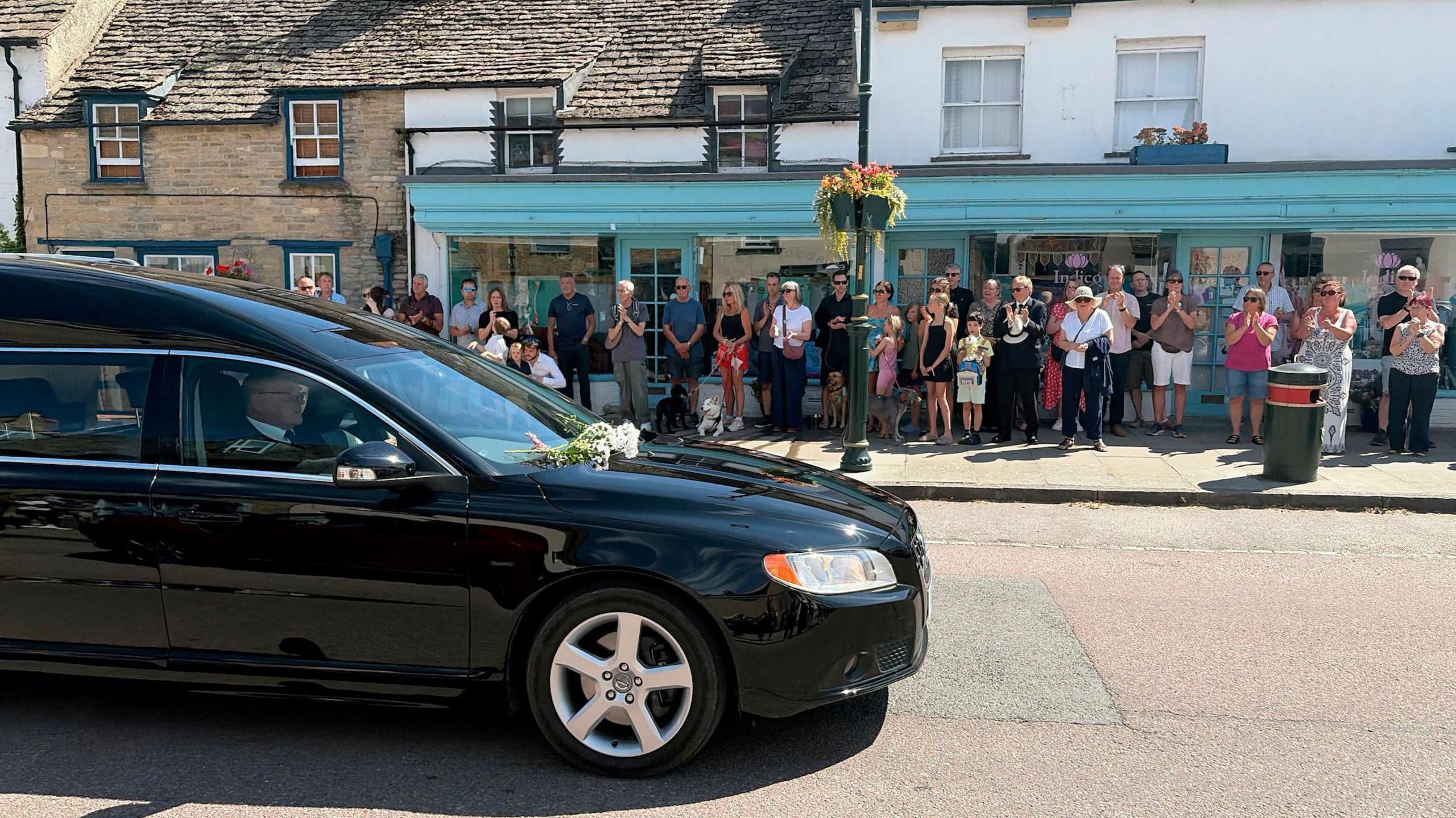 A black hearse in a historic street on a sunny day. People line up along the pavement.