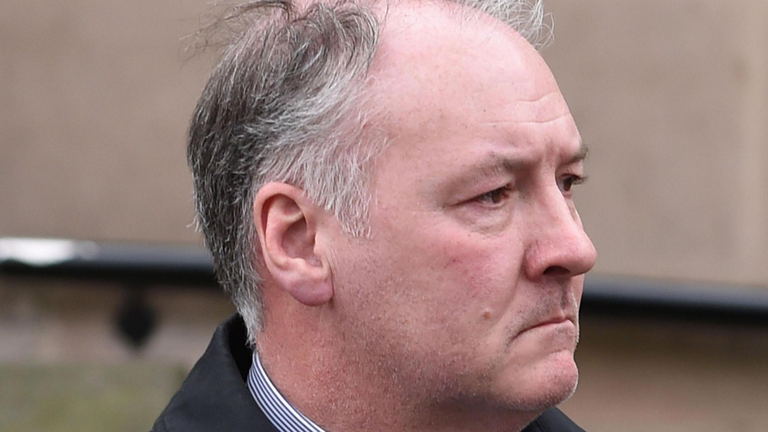 A balding man with his face angled away from the camera and a serious expression. He is outside and his grey and white hair is blowing in the wind.