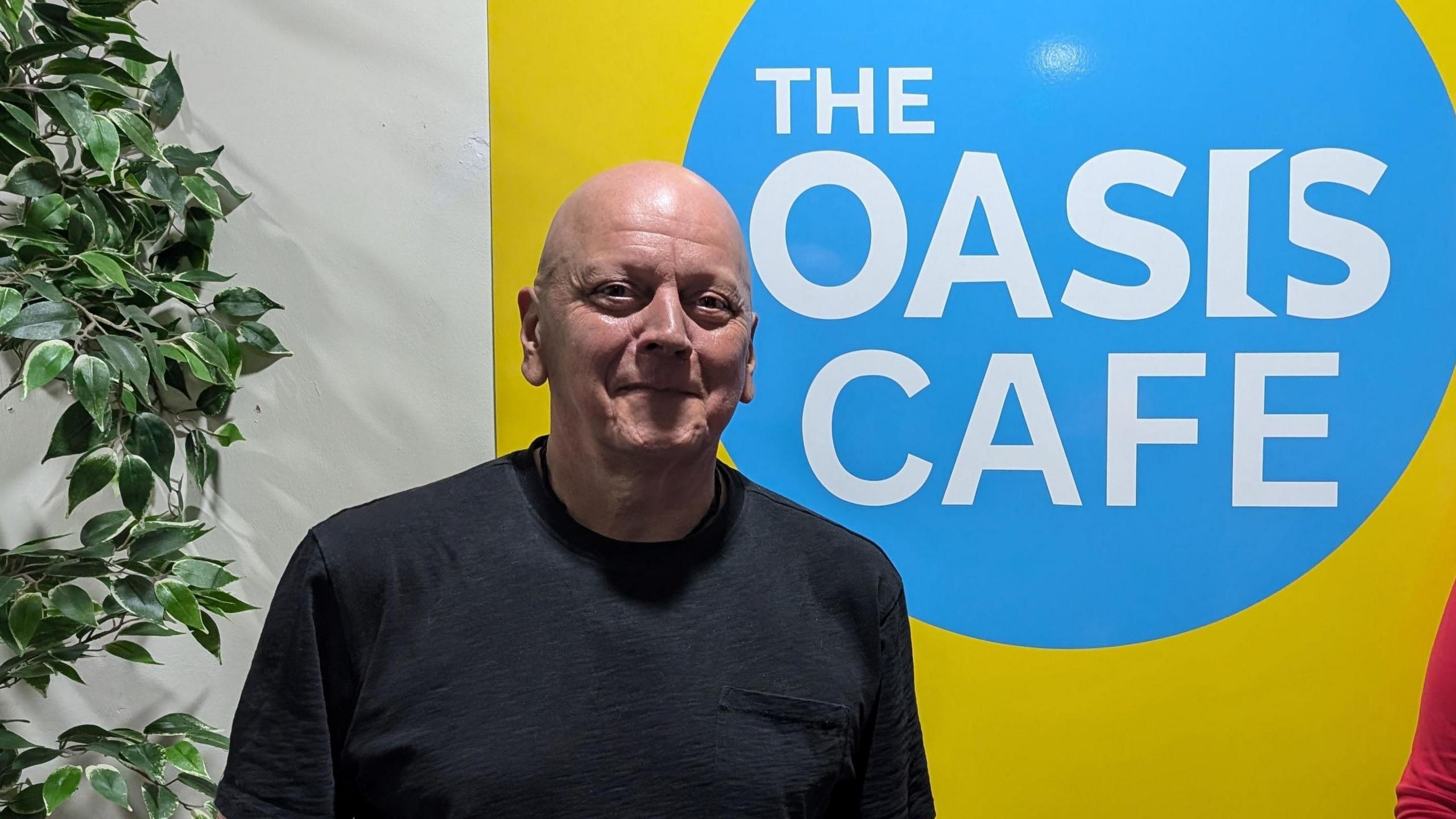 Colin McGregor-Paterson smiles directly at the camera and is wearing a black T-shirt. He is standing in front of a blue and yellow sign which reads "The Oasis Cafe".