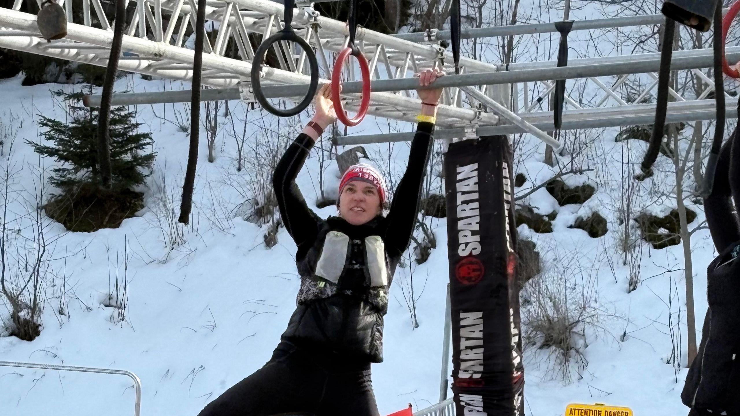 Fran Bullock pulling herself across monkey bars. She is wearing a hat and warm clothing. There is snow in the background.