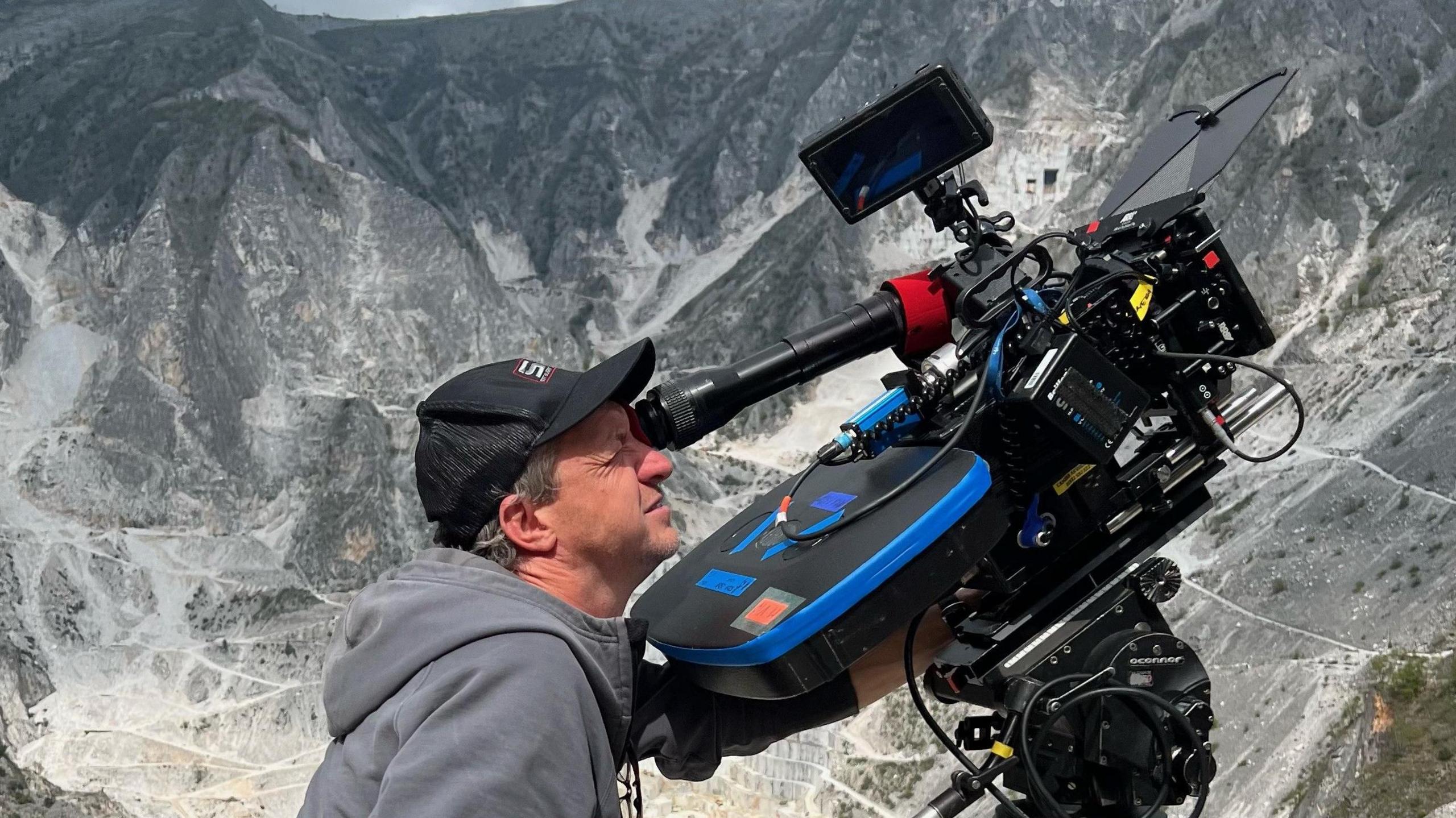 Lol Crawley, who has short grey hair and is wearing a black baseball cap and grey hoody, looking through the lese of a large black camera which is pointed upwards, at something off to the right of the picture. In the background are huge grey mountains.