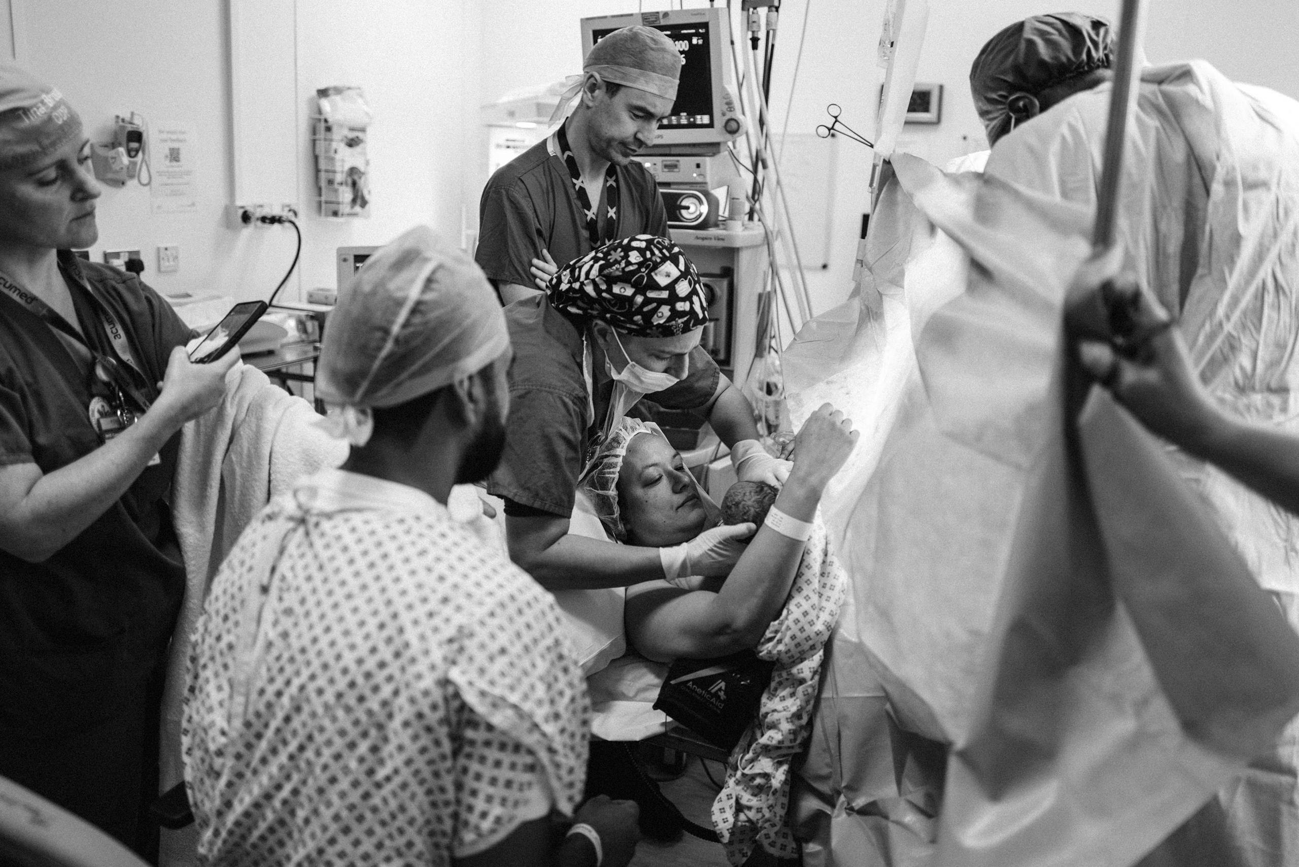 A mother holding her newborn moments after delivery, assisted by healthcare staff in scrubs. Her partner sits close by, and a staff member captures the moment on a phone.