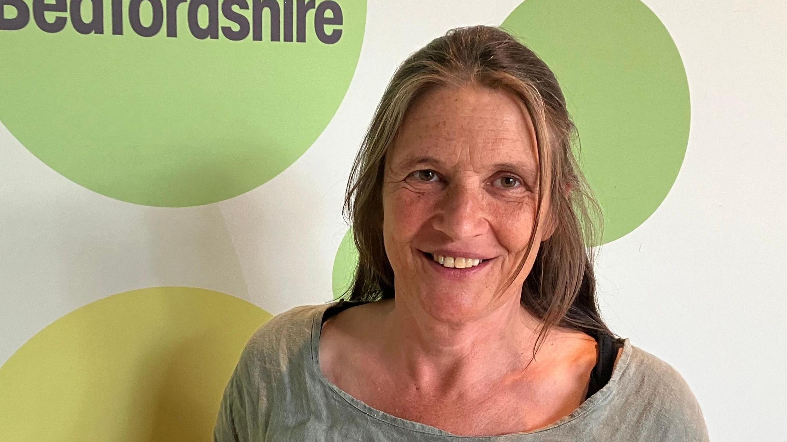 A picture of a woman standing in front of a wall that has circles on it and the word Bedfordshire. She has a greyish top on and has her brown hair framing her face.