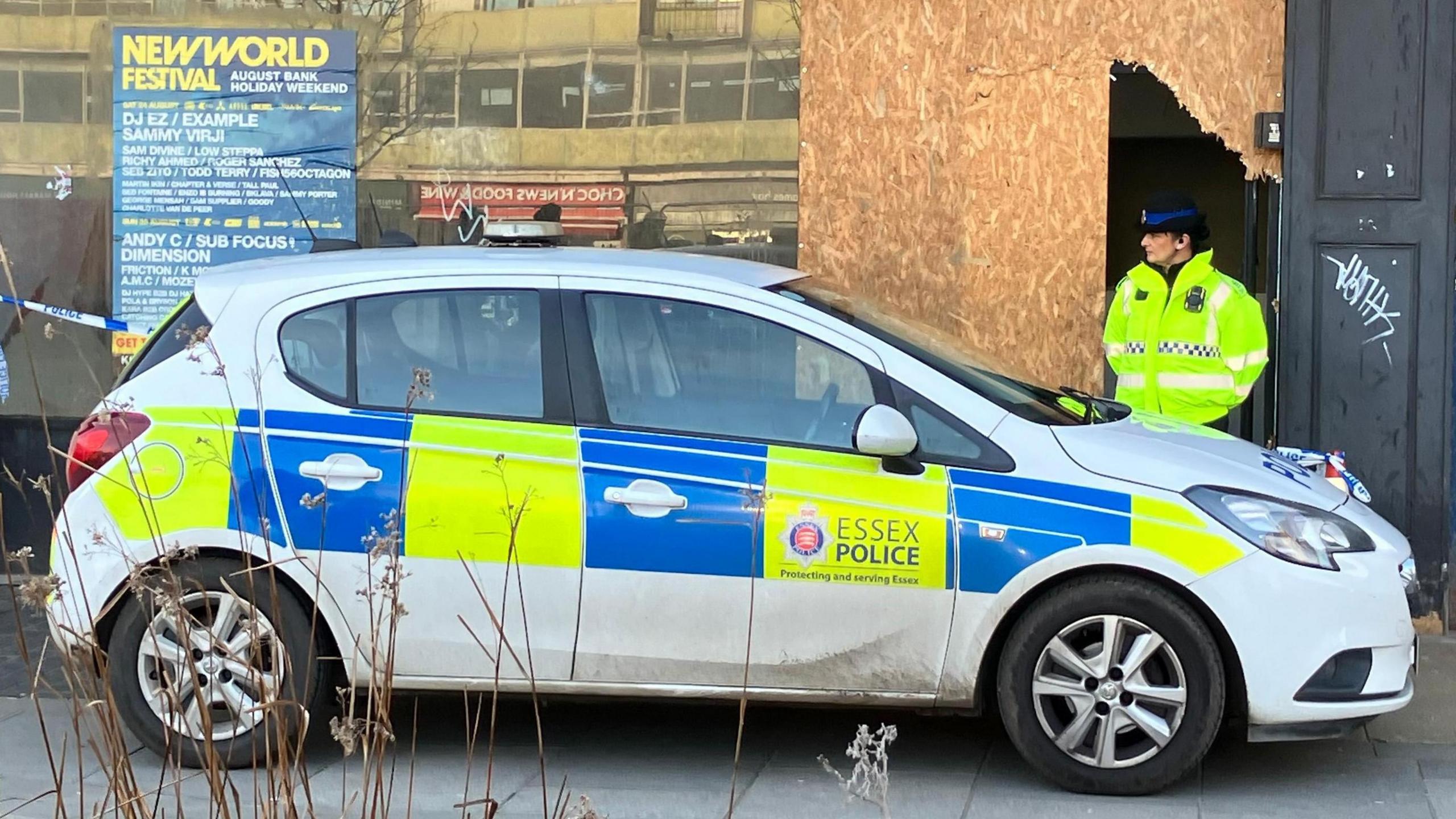 A female police officer wears a hi-vis jacket and black hat as she guards a vacant shop, which has been partially boarded up. She stands behind a hatchback police car and next to blue and white tape.