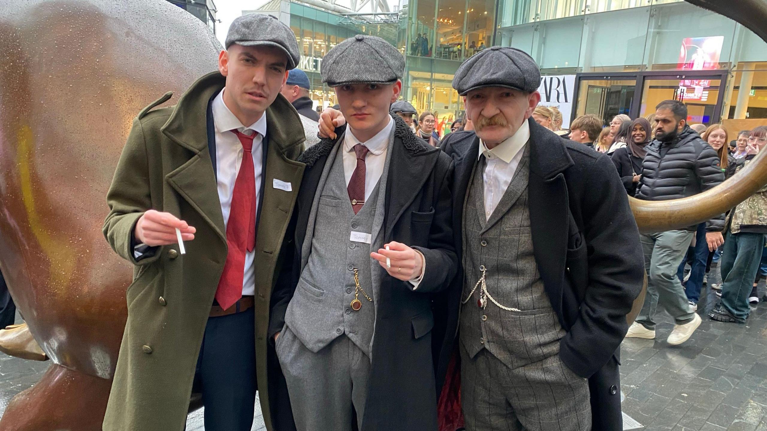 Three men in grey flat caps and overcoats. two of them are holding cigarettes 