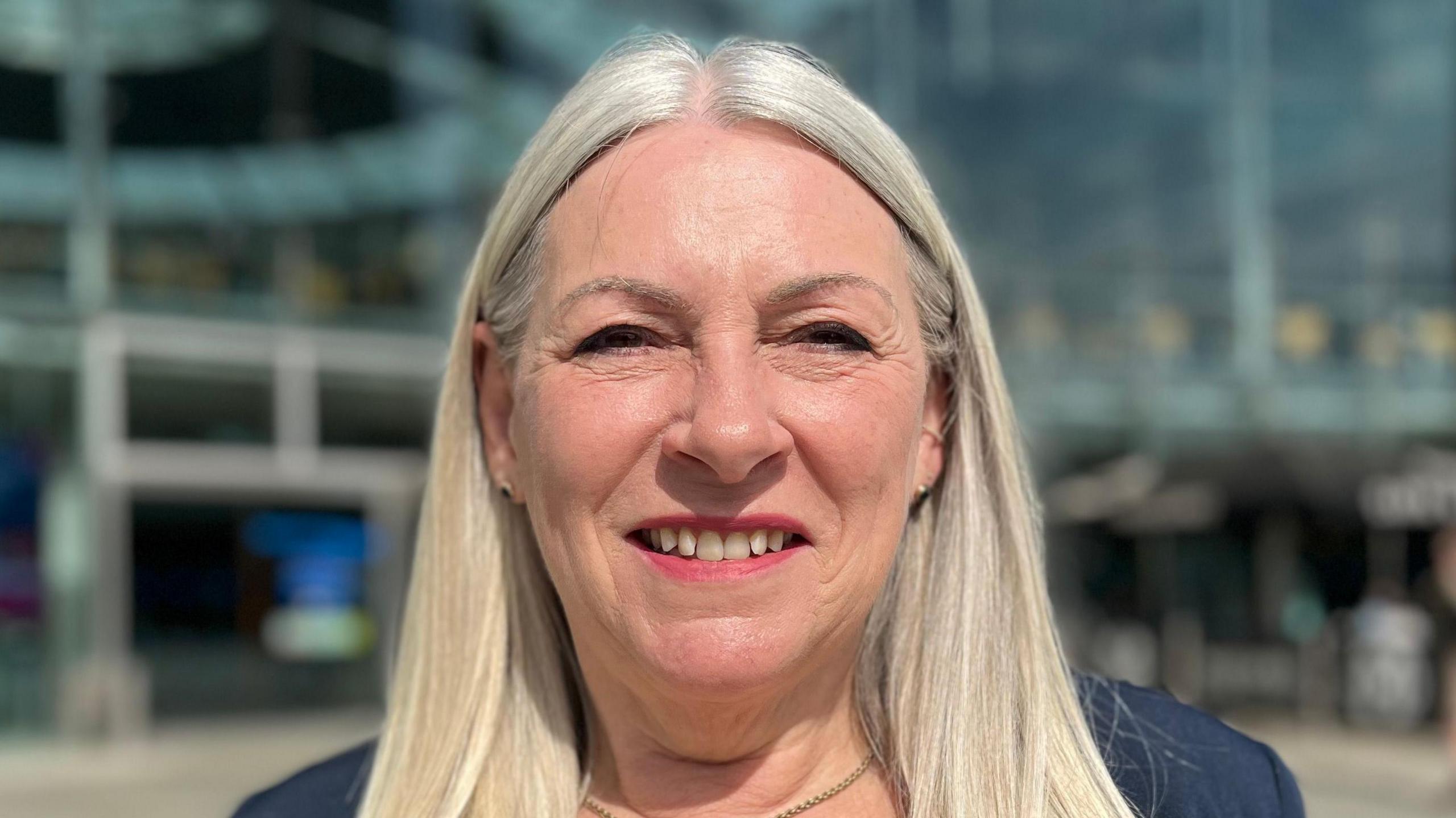 Kay Mason Billig smiling and wearing a dark blue suit jacket. It is clearly sunny weather. She is standing in front of the Norwich Forum building.