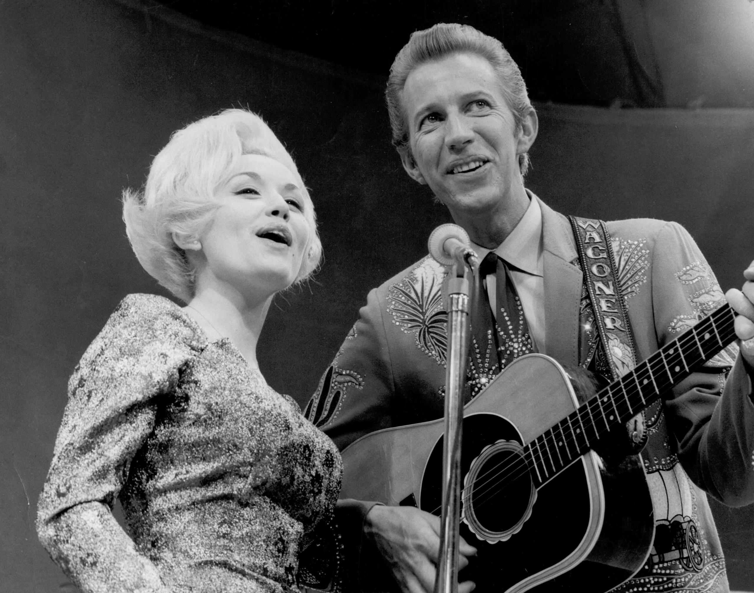 Dolly Parton and her collaborator Porter Wagoner perform onstage in circa 1967. She has a blond bob and a patterned dress and sings into the microphone with Porter, who plays guitar and is in a flashy suit and tie.