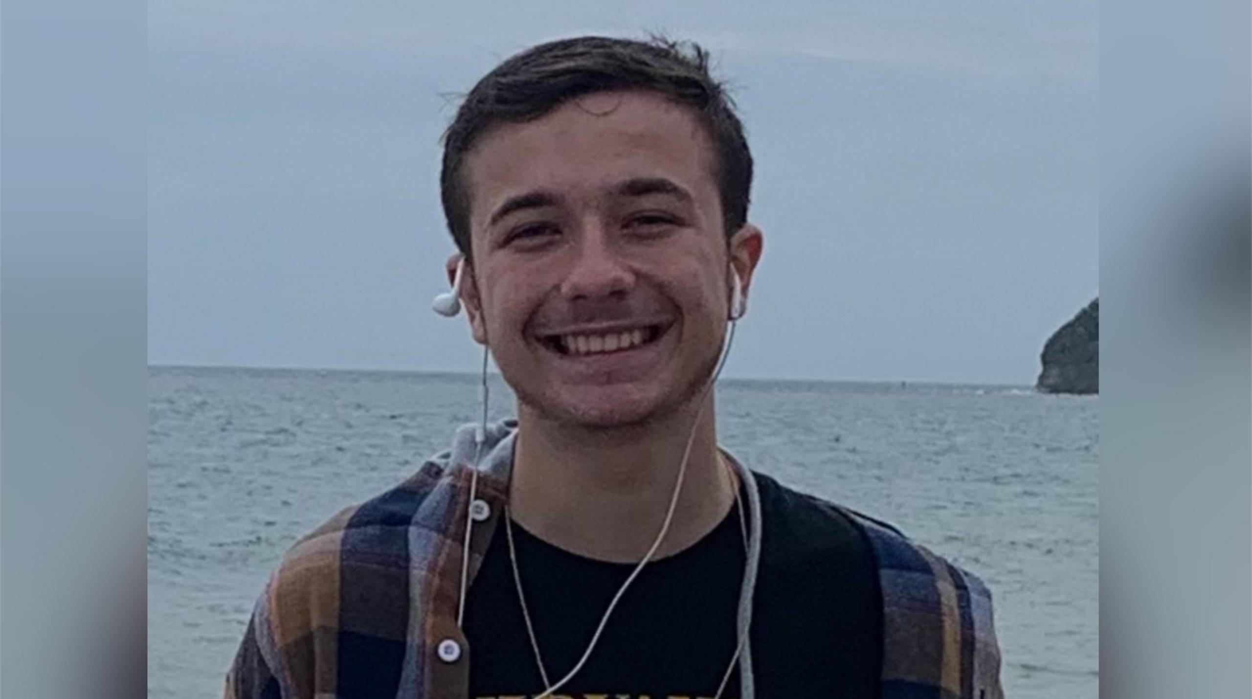 Aidan Webb with short dark hair smiling at the camera with the sea behind him.  He has earplugs around his ears and is wearing a multicoloured striped shirt.