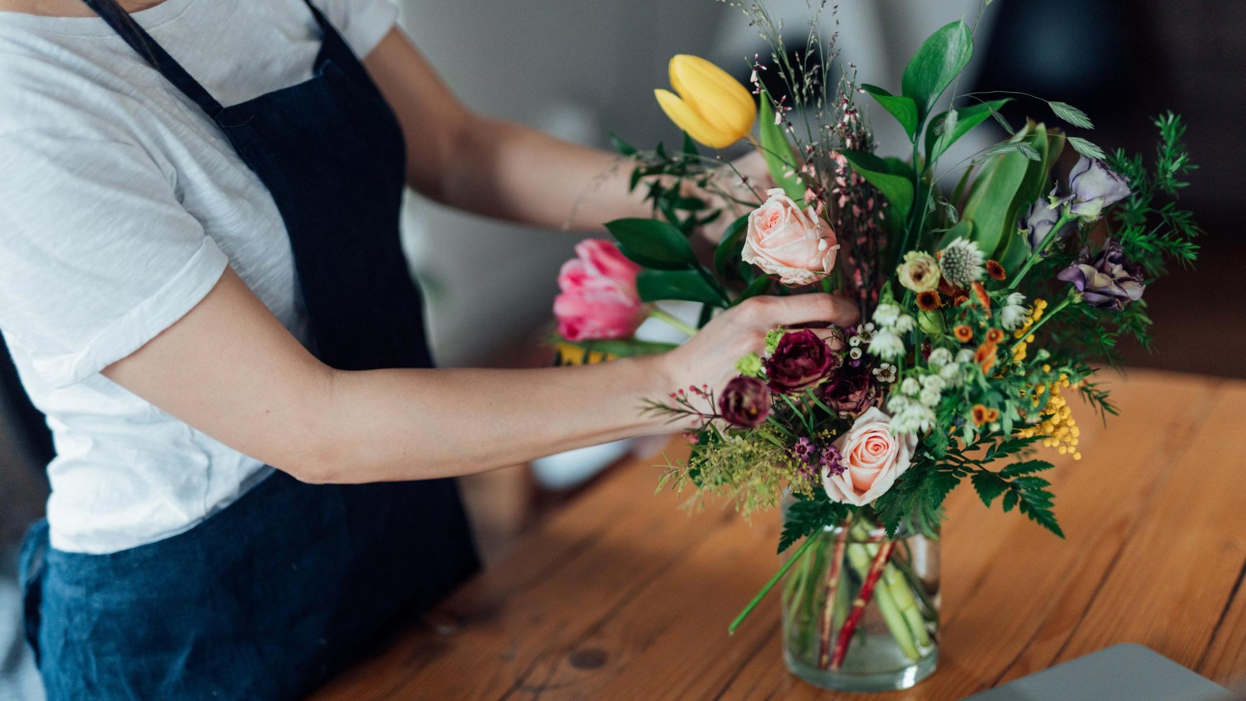 Person in a white shirt and dark blue apron arranging a colourful bouquet of tulips, roses, lisianthus, and foliage in a glass vase on a wooden table