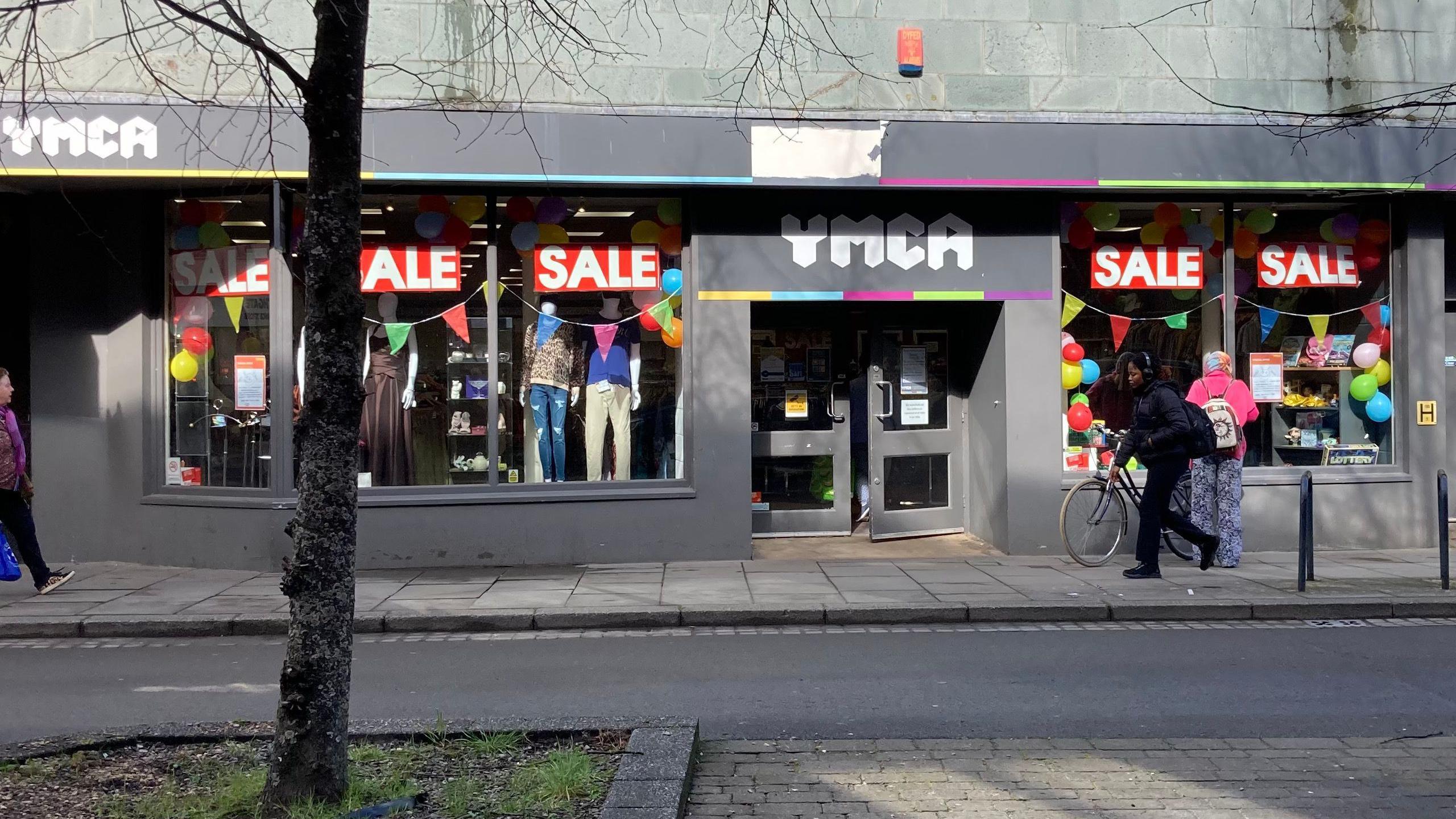 A photo showing a YMCA charity shop in Gloucester with a front door, windows and a tree outside