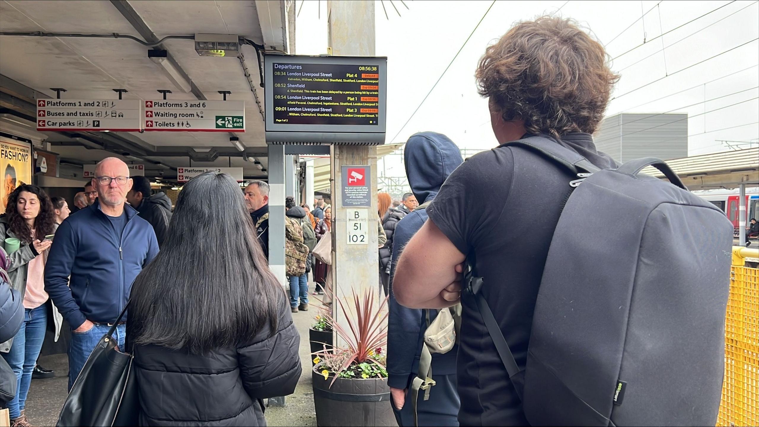 People standing on a busy platform at Colchester railway station. Most people in the picture have their backs to the camera and are looking at a travel information screen, which shows all journeys being delayed. It is a grey day and most people are wearing coats or jumpers.