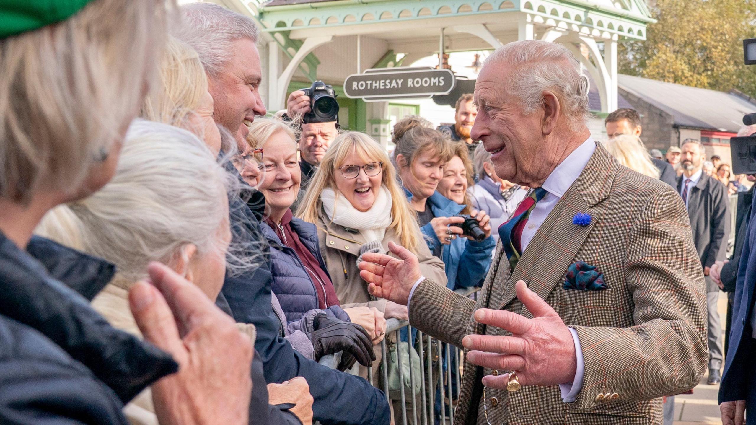 King Charles III, who is wearing a brown check tweed jacket and waistcoat with a colourful tie and pocket handkerchief, smiles with his arms outstretched as he meets members of the public during a visit to mark the 150th anniversary of The Albert Hall in Ballater, Aberdeenshire. A railway style sign reading Rothesay Rooms hangs from the wooden arch of the former Royal Railway Station above the small crowd.