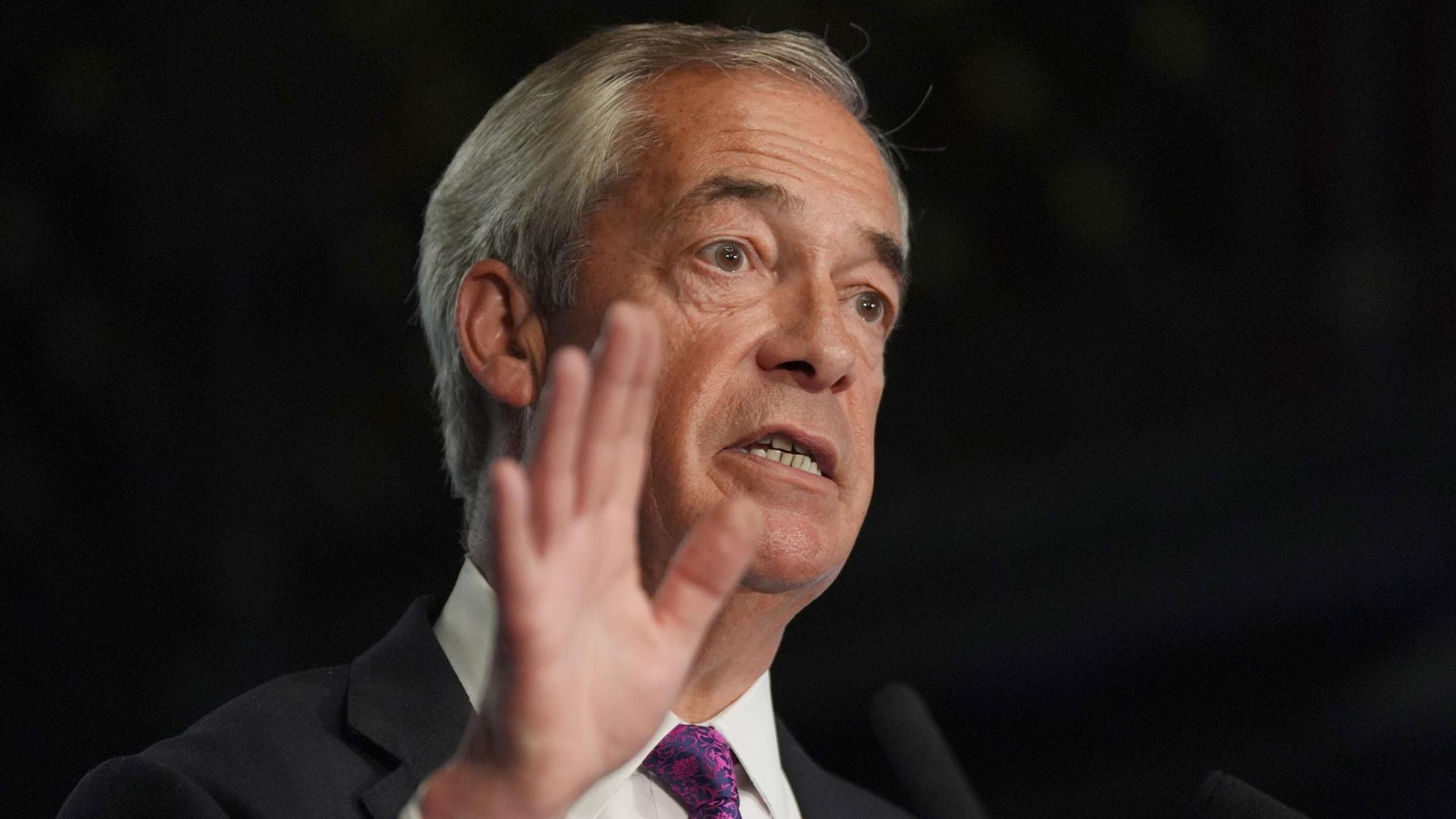 A man wearing a suit and tie with his hand in the air. He has white hair 