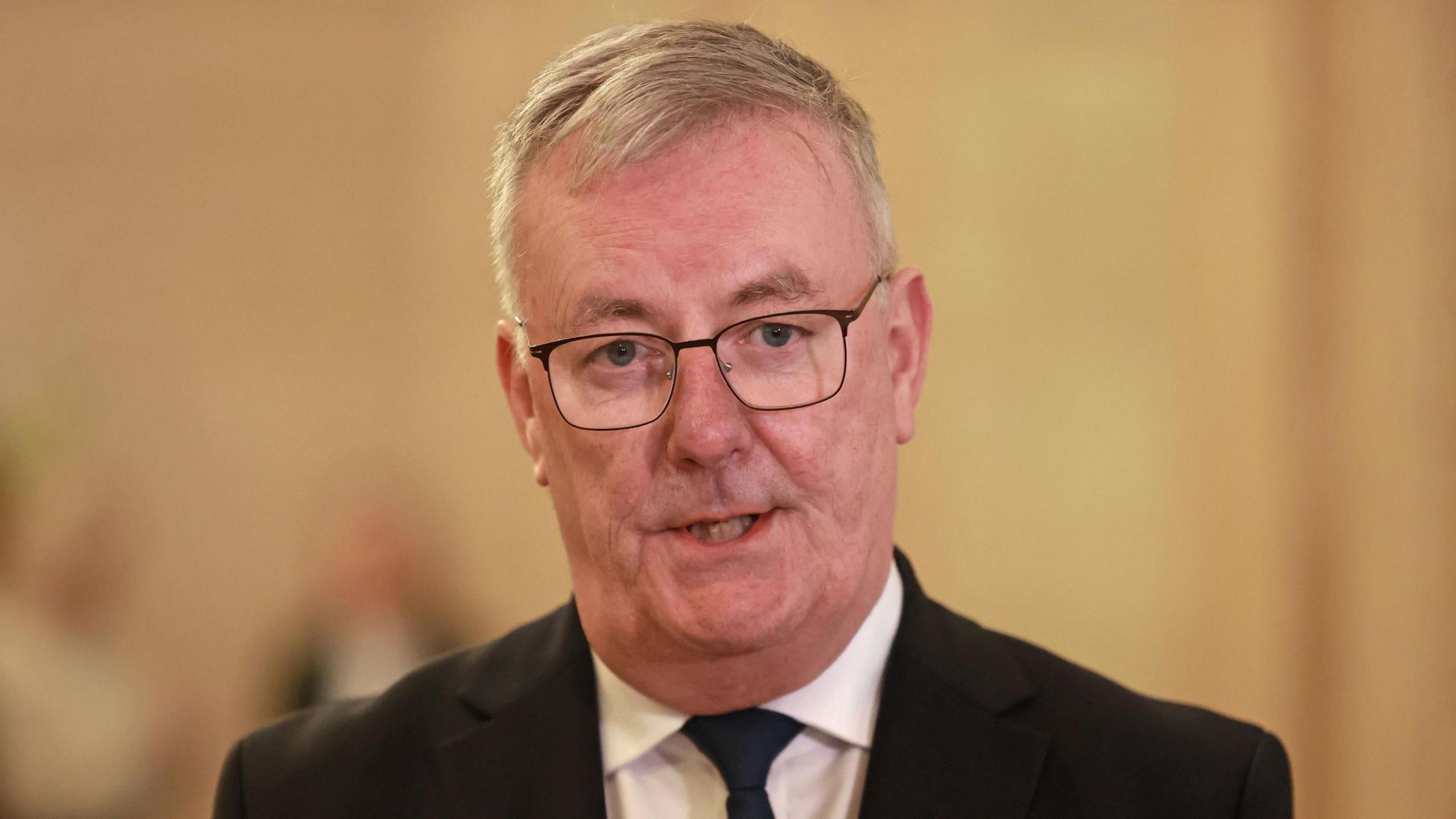 Mike Nesbitt wears a black suit, white shirt and black tie. He has short grey hair and clear lensed spectacles. Behind him a cream/yellow wall. 