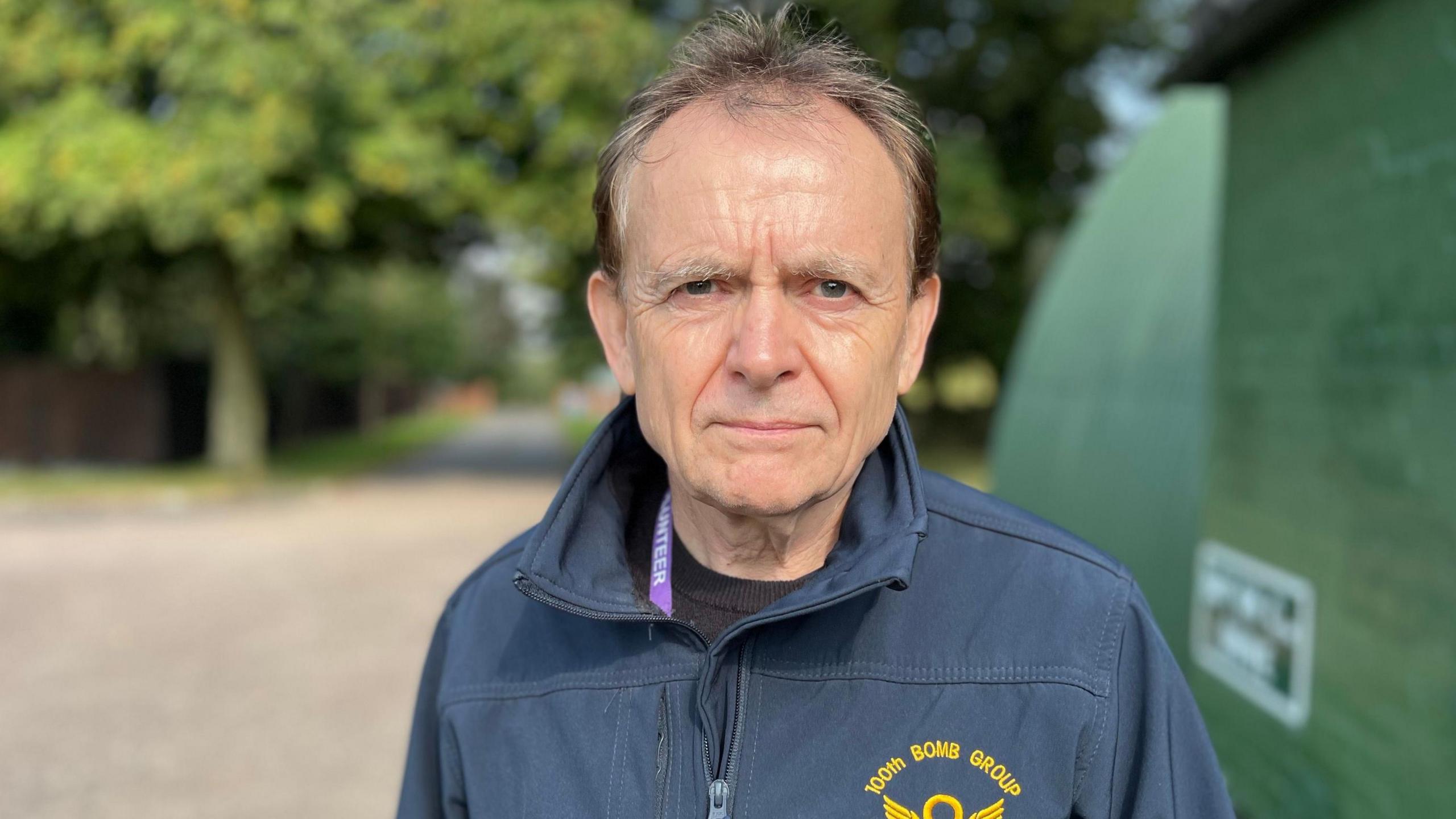 Reg Wilson wearing a blue jacket with "100th Bomb Group" written on the front standing outside a military building with trees in the distance.