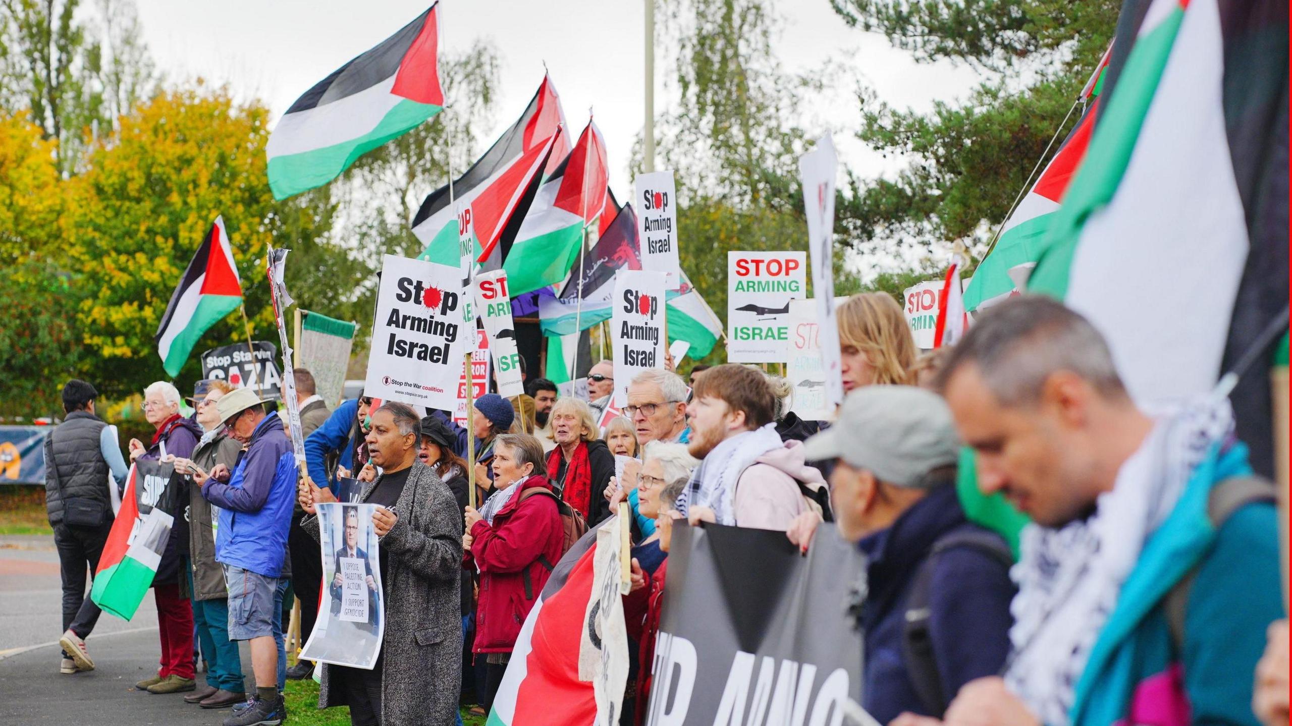 A large group of protesters holding Palestine flags, banners, and signs with slogans such as "Stop Arming Israel" stand in a line with trees in the background