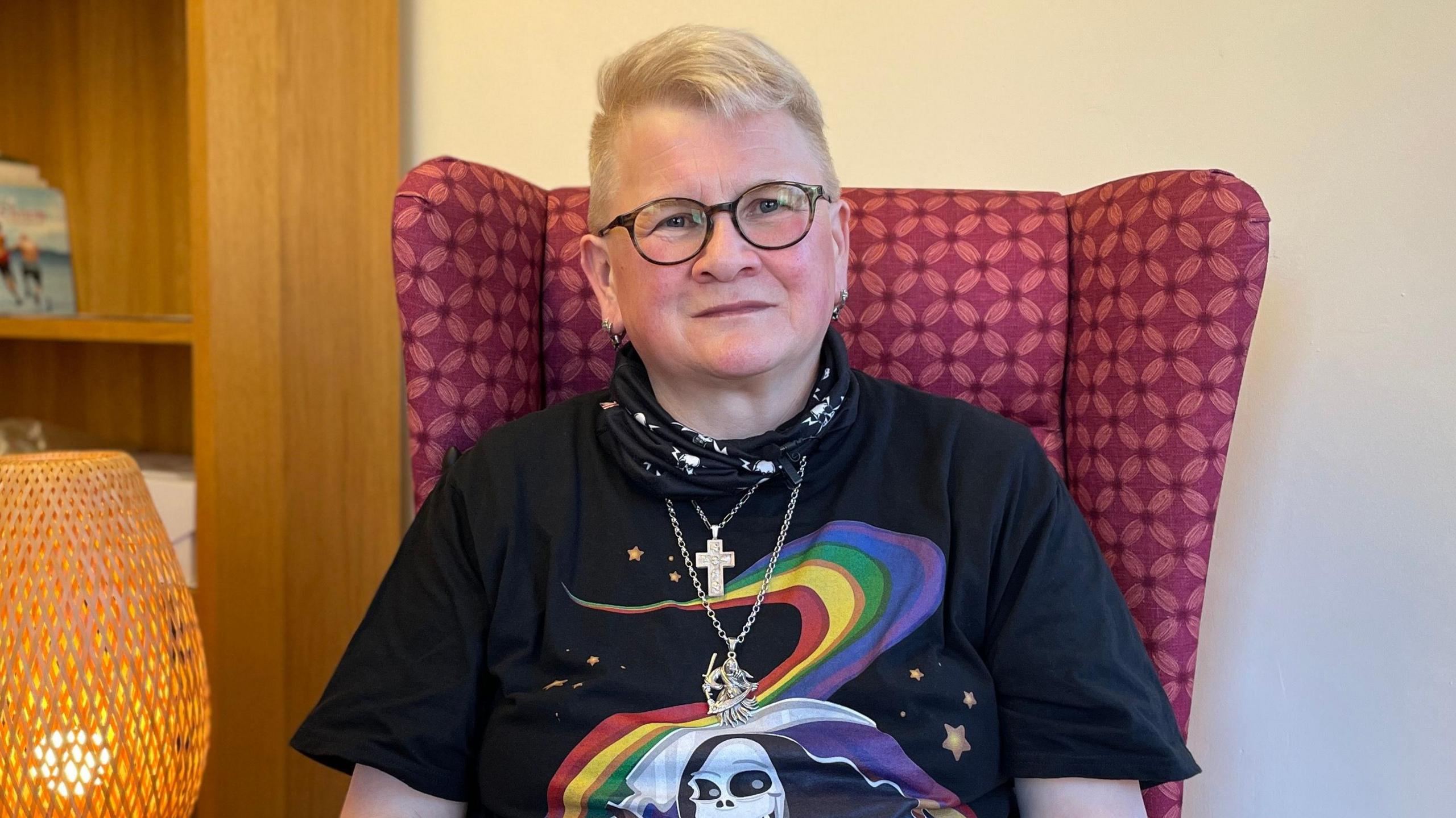 A woman with short blonde hair sits in a red patterned armchair. She is wearing dark-framed round glasses. She is wearing two silver pendants and a neckerchief. She is wearing a black T-shirt with a rainbow across it and a cartoon of the grim reaper on it. In the background there is a bookcase.