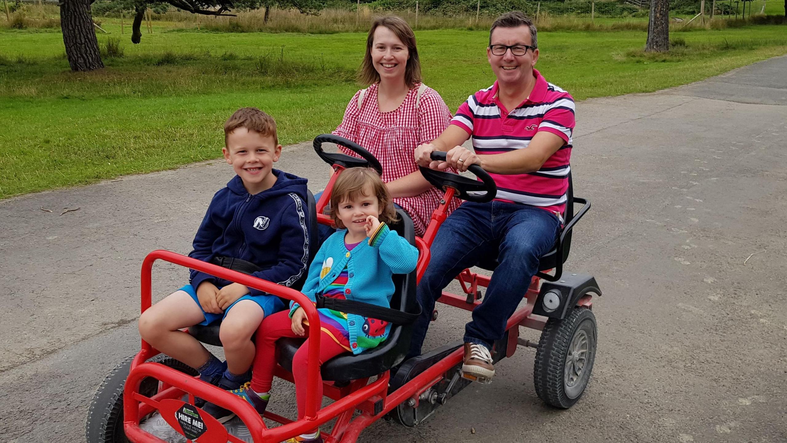 Nicola, Darren and their two children on a pedal-powered cart red cart. Nicola is wearing a red gingham dress and Darren is wearing a pink and white striped polo shirt, jeans and trainers. Ffion is wearing a blue cardigan and has her finger in her mouth. Oscar is wearing a navy hoody and shorts. All are smiling at the camera. 