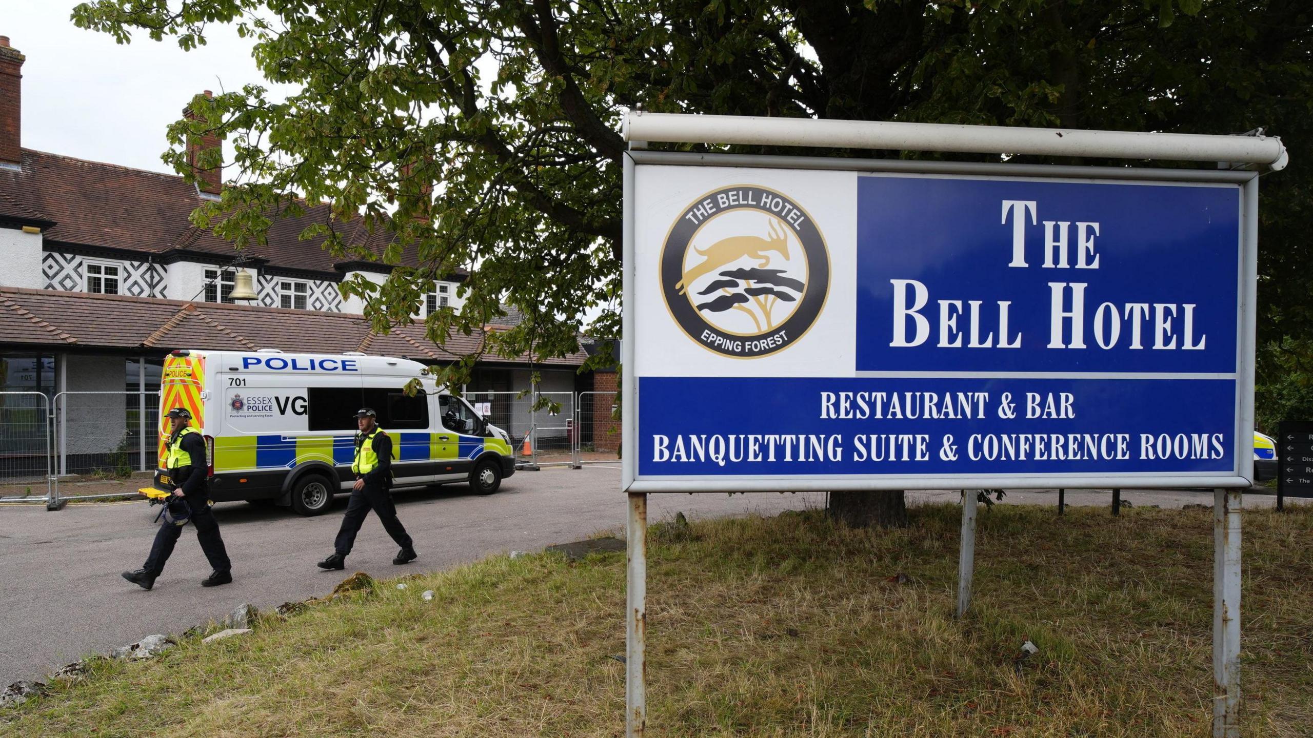 Two police officers walking next to the hotel, which is surrounded by a thin metal fence. A police van has also parked next to the building. In the foreground is a large blue sign which reads 'The Bell Hotel'.