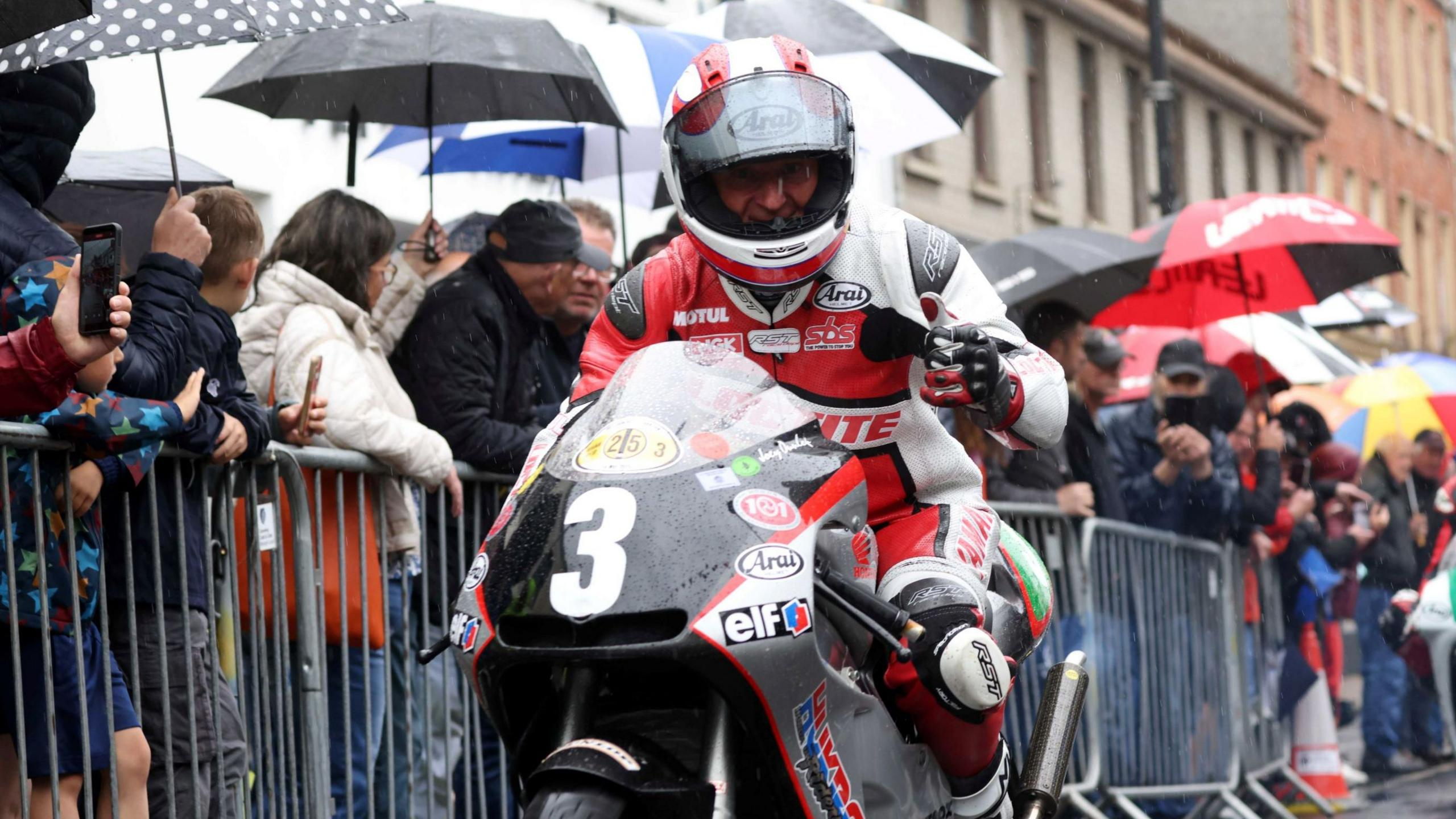 A motorbike rider, dressed in red and white protective gear, is on a racing bike with the number 3 on the front. He has a helmet on but you can tell he is smiling at the camera. There are rows of people behind him in the street. They are separated by grey metal dividers. A number of the spectators have umbrellas up.