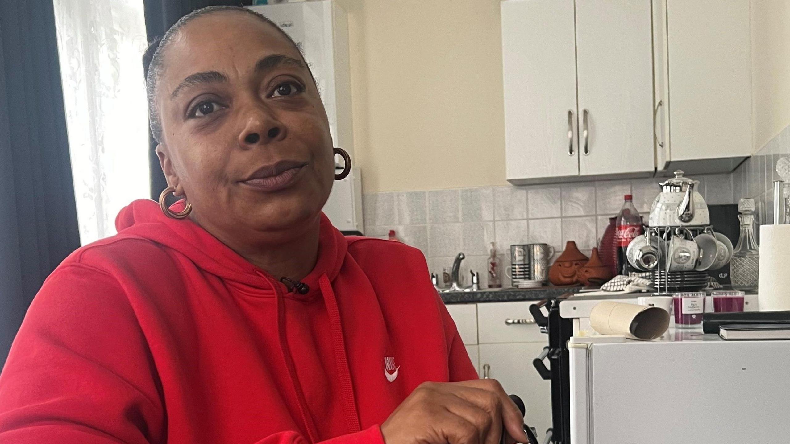 Deborah Morgan, wearing a red hooded sweatshirt and holding walking stick, sitting in her kitchen