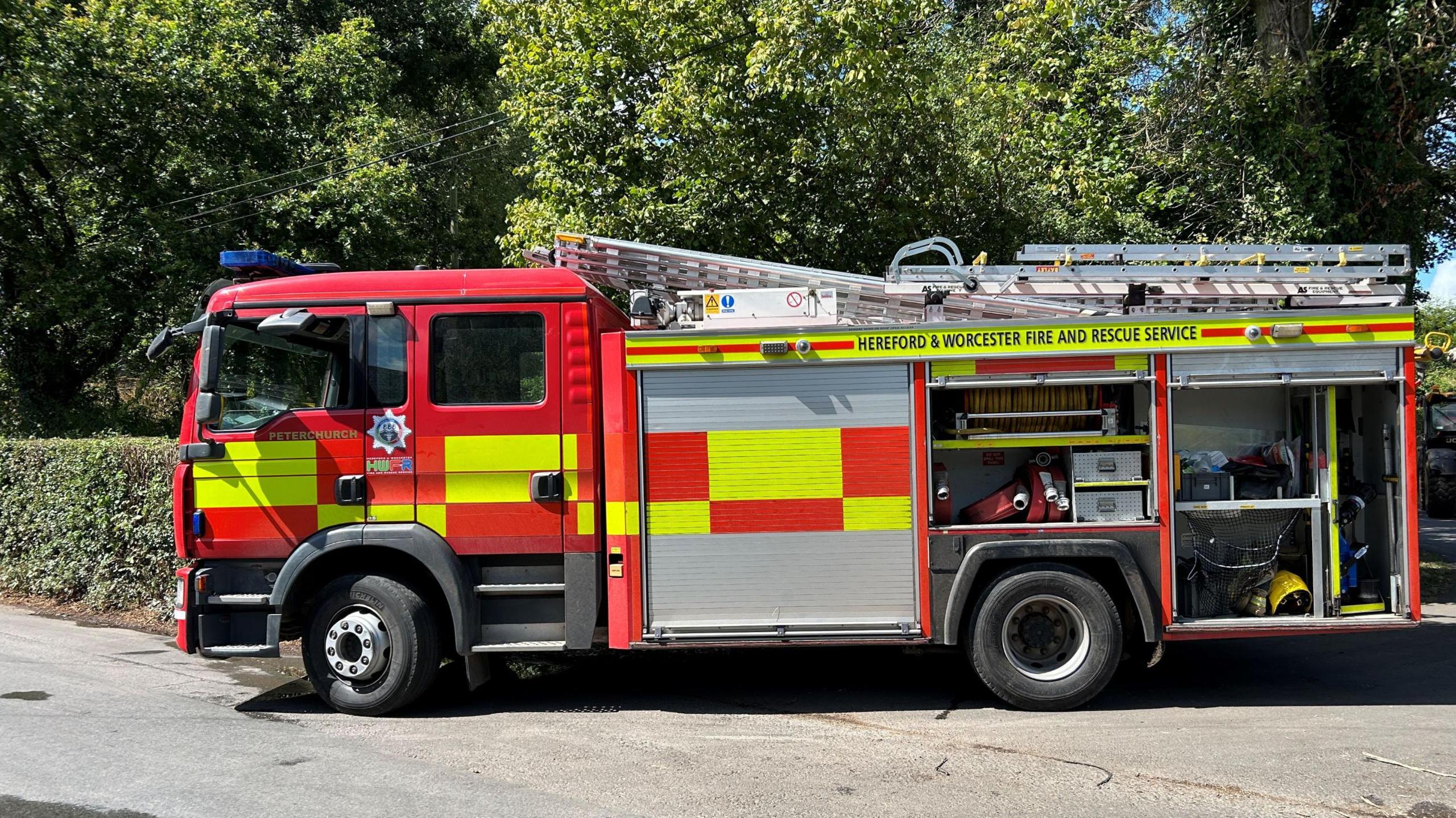A fire engine in a countryside lane. The engine is mainly red with yellow rectangles on the side. A ladder is on the roof.