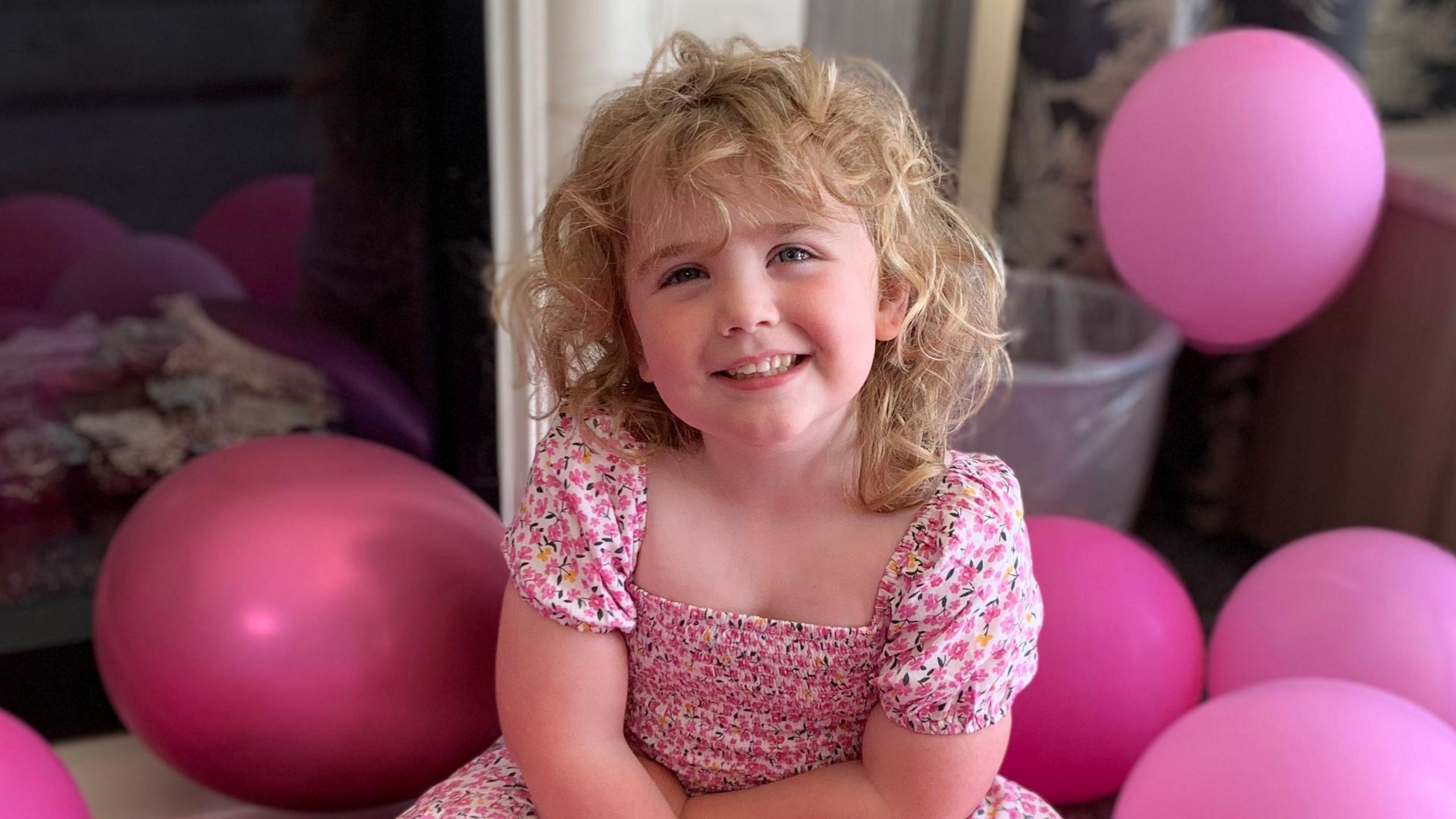 A five-year-old girl in a pink dress smiles into the camera surrounded by pink balloons. 