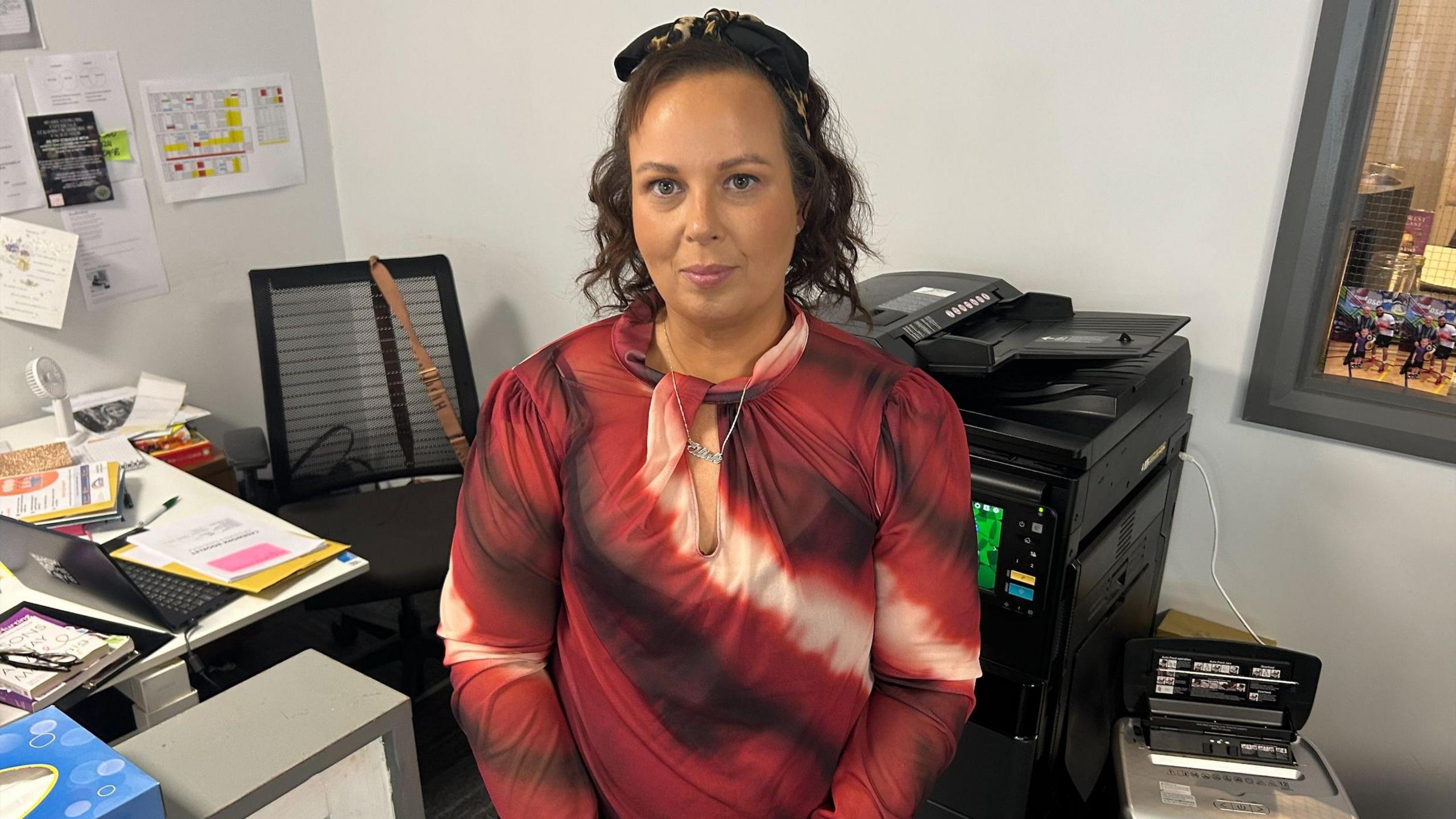 A woman is sitting in a small office. There is a printer and laptop beside here. Paper, a pen and glasses sit on a table. The woman is wearing a red patterned dress. 