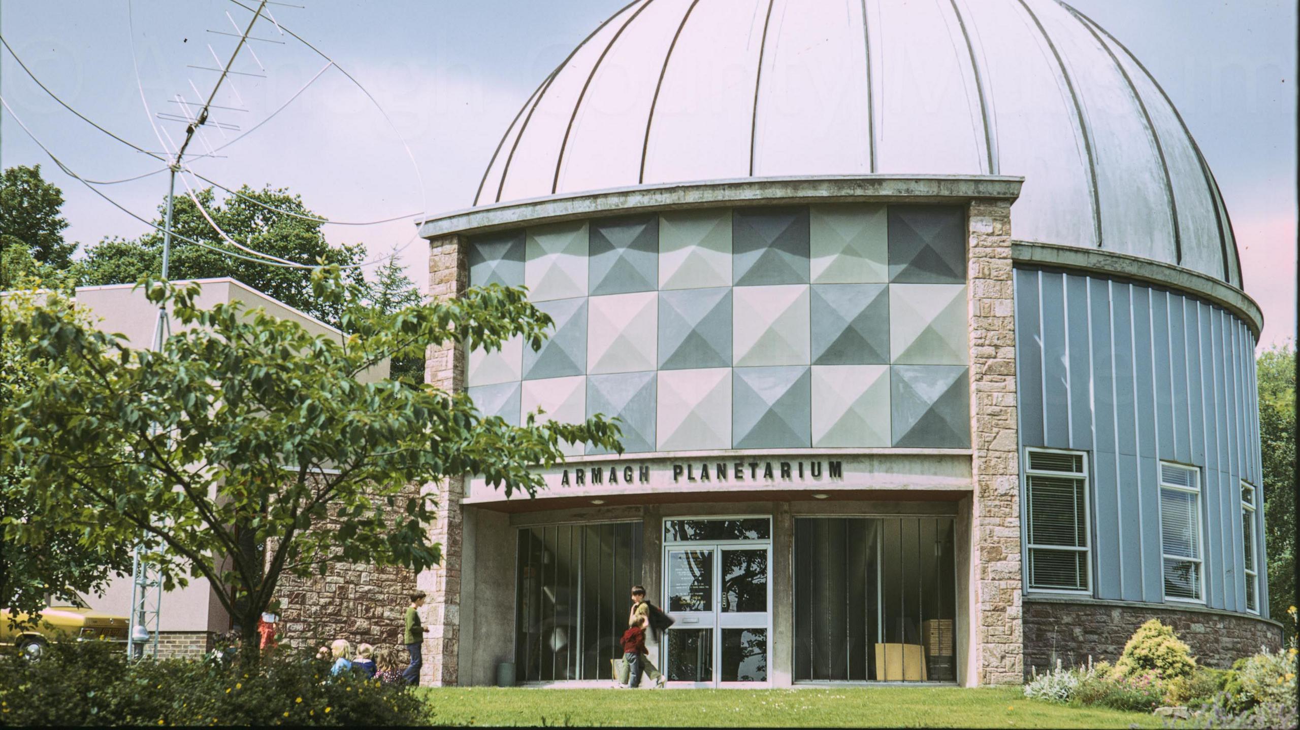 A digitalised photo from a film of the dome shaped planetarium. View of the front of the planetarium with 'Hall of Astronomy' visible through trees on the left above which is the dish of the radio telescope. c1973