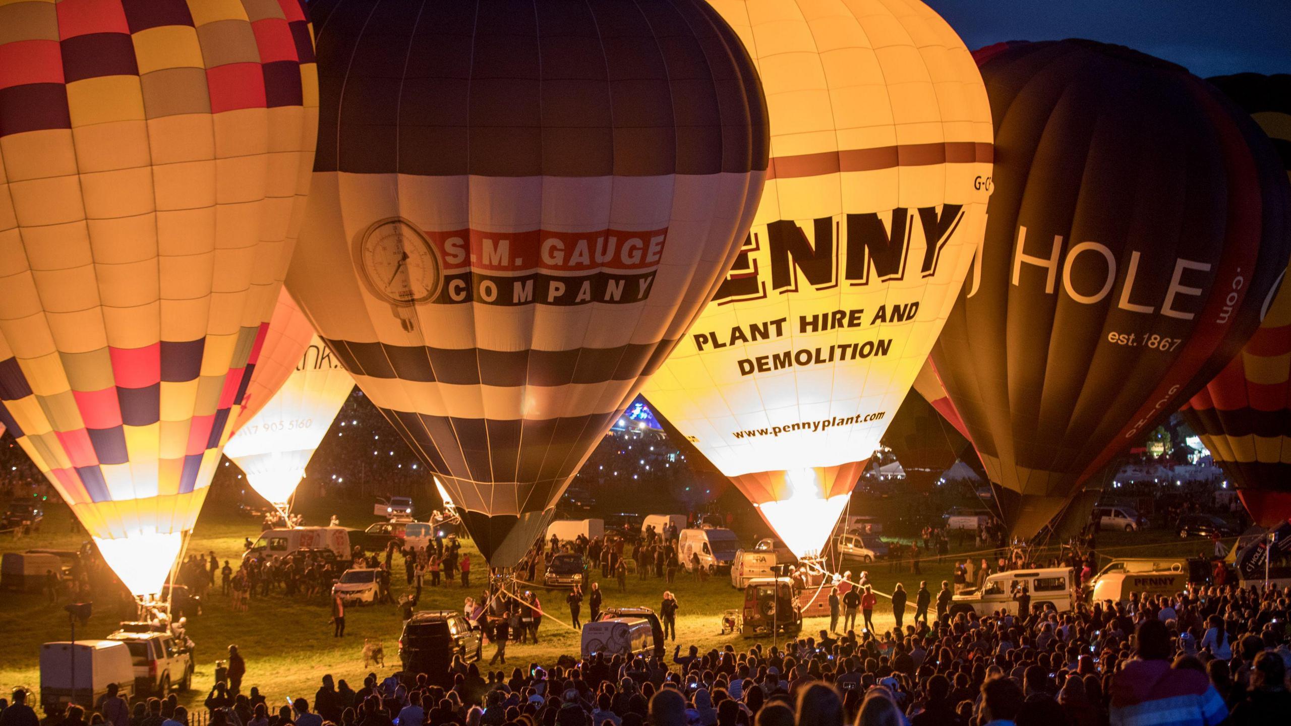 Several hot air balloons that are inflated but tethered to the ground. A few of them have fire inside. A large crowd of people can be seen watching the display. 